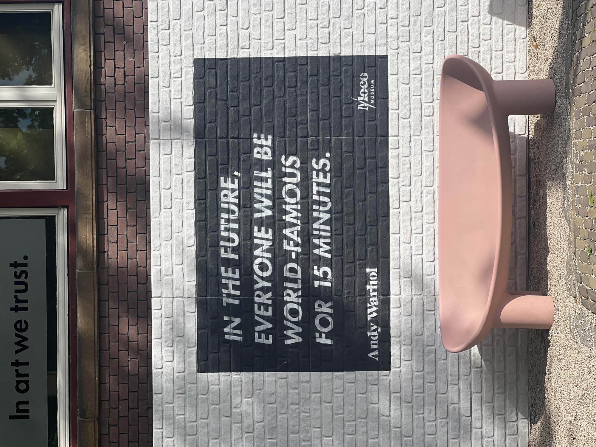 The image shows a wall with a mural that reads "In the future, everyone will be world-famous for 15 minutes" by Andy Warhol. Below the mural, there is a pink bench. The wall is painted white bricks, with a section painted black featuring the mural. The text "Moco Museum" is also visible on the wall. The scene appears to be outdoors, with gravel on the ground. The building above the wall has windows reflecting trees and sky. This is likely an exterior wall of the Moco Museum in Amsterdam, Netherlands, known for its exhibitions of modern and contemporary art.