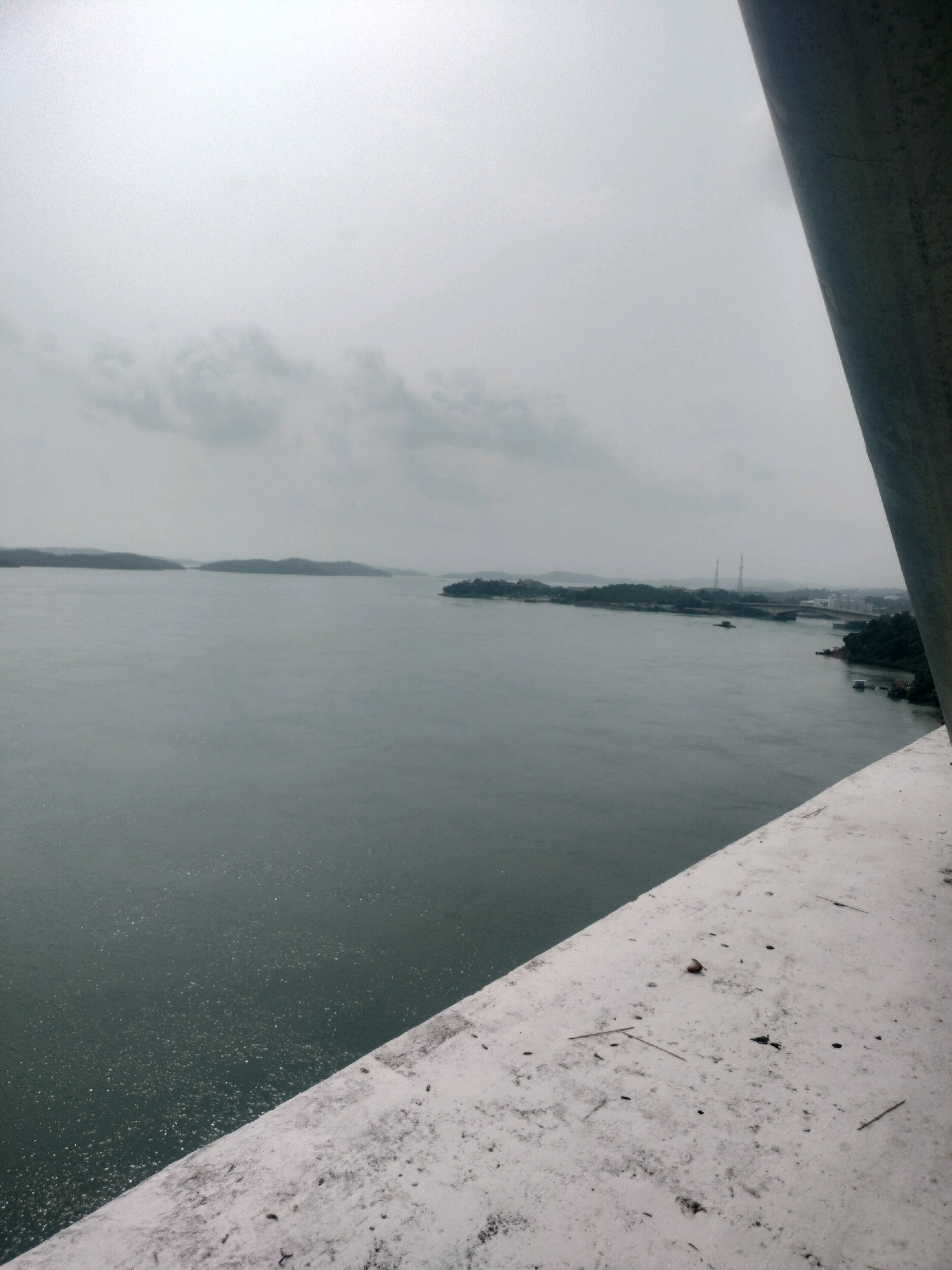 This is an eye-level, medium shot of a vast body of water, possibly a strait or bay, under an overcast sky. The water appears dark green and is slightly rippled, with a scattering of bright reflections suggesting sunlight breaking through the clouds or a textured surface. In the distance, a low-lying, tree-covered island or landmass stretches across the horizon, dotted with structures and what appear to be transmission towers. A bridge is visible in the middle distance, connecting two points of land.
The foreground is dominated by a concrete structure, likely a bridge railing or walkway, with a rough, textured surface. The concrete is speckled with dark debris, possibly dirt, leaves, or small stones. The perspective suggests the photo was taken from a high vantage point, looking out over the water and towards the distant shore. The overall mood is muted and somewhat somber due to the cloudy weather.
This scene is likely the Batam Outer Ring Road (BORR) crossing, a major infrastructure project in Batam, Indonesia. The specific location could be along the stretch of the BORR that spans over the waters connecting different islands or peninsulas of Batam.