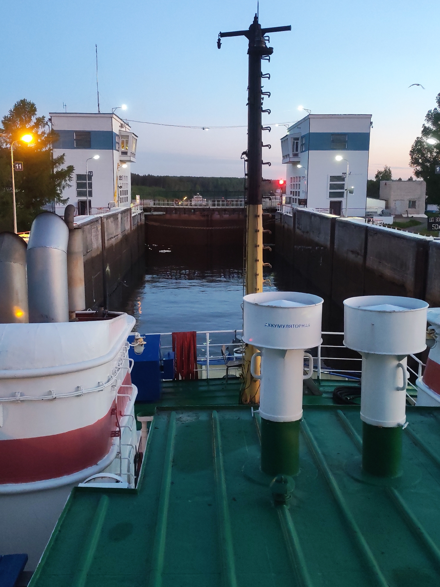 The image depicts a ship positioned within a concrete lock chamber during twilight or early morning. The sky is a soft blue and pink, suggesting dusk or dawn, with streetlights and lights from buildings already illuminated.
In the foreground, the green, ribbed deck of a ship is visible. On the left, a white and red lifeboat or tender is mounted. Further right, three prominent silver exhaust pipes rise, and two large, white cylindrical structures with green bases stand on the deck. One of these cylinders clearly displays the Cyrillic text "АККУМУЛЯТОРНАЯ" (AKKUMULYATORNAYA), meaning "BATTERY ROOM" or "ACCUMULATOR ROOM." A tall, dark pole, equipped with numerous cleats or hooks, stands centrally on the deck, extending upwards.
The ship is contained within a calm, dark-watered lock. On both sides of the lock, modern, white control buildings with distinctive blue horizontal stripes are visible. These buildings feature multiple windows and are illuminated by external lamps and internal lights. On the left building, a number "12" is visible on the wall, and "11" is on a nearby streetlight pole. The right control building displays a prominent red signal light, indicating a 'stop' or 'do not proceed' instruction. Faint Cyrillic text, including "ДО 300" (DO 300) and possibly "50," is discernible on a sign near this building.
In the background, a distant tree line and what appears to be a bridge or embankment stretch across the horizon. A lone bird, possibly a seagull, is visible flying in the sky above the right control building. The scene conveys a quiet and serene atmosphere of maritime activity, likely waiting for the lock to fill or empty.