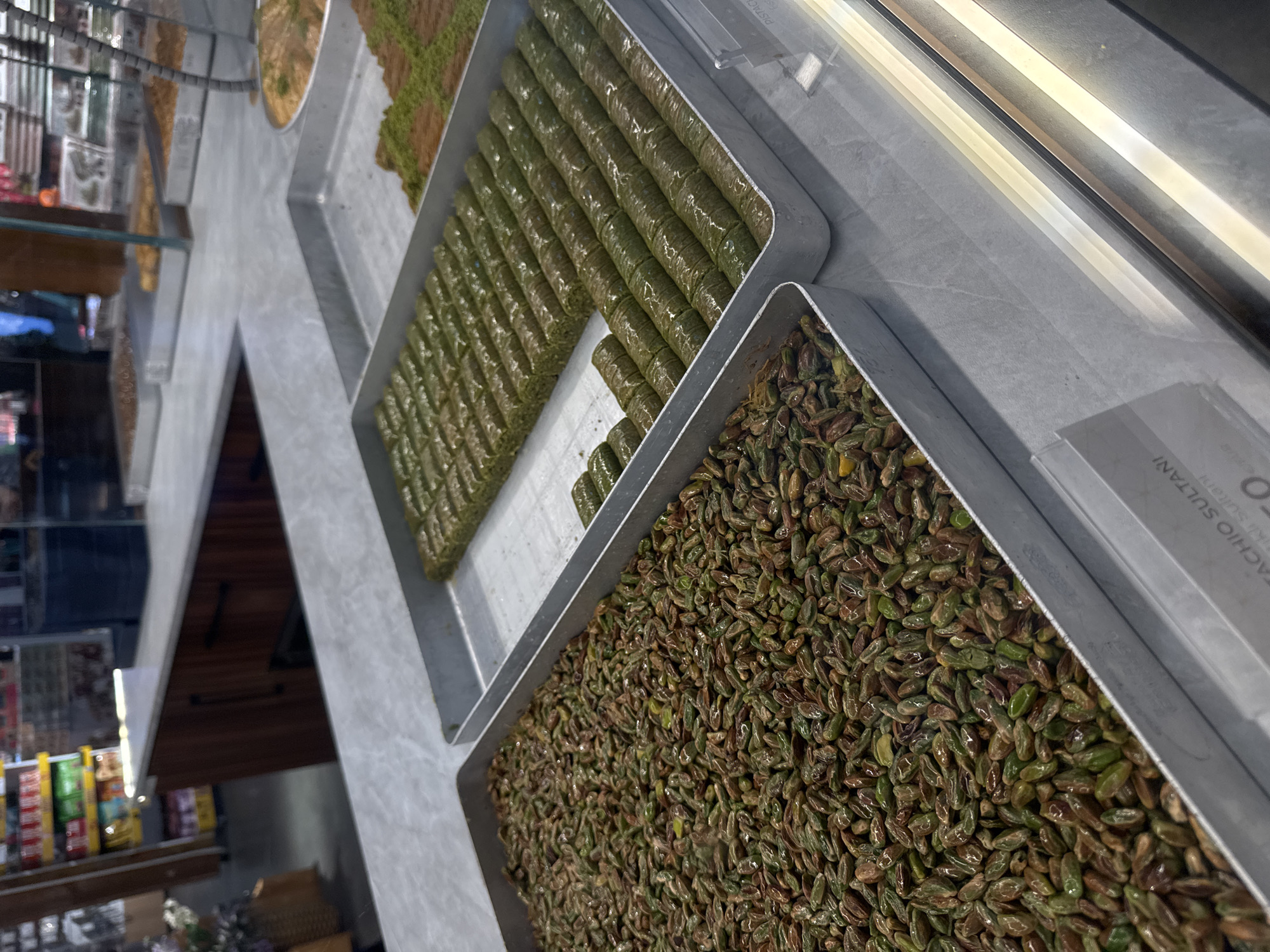 The image shows a display of Turkish pastries and ingredients in a pastry shop, likely located in Arnavutköy, Turkey. Three trays are visible, filled with different types of pistachio-based desserts. The closest tray is filled with loose pistachios. The tray further away from the viewer contains rolled pastries, likely a type of baklava, and the farthest tray contains another variety of layered pastries. The display is within a glass case, and the shop's interior is visible in the background, showing other goods and a shelving unit. A sign identifying "Pistachio Sultani" is visible.