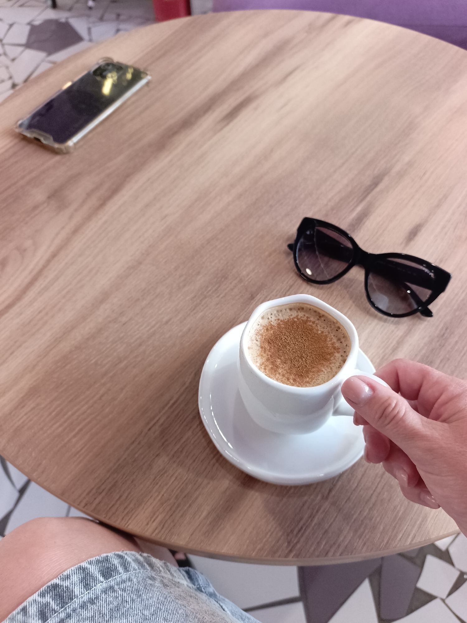 A person's hand is holding a white cup of coffee with cinnamon on top. The coffee cup rests on a white saucer, which is on a light brown wooden table. Next to the coffee cup are a pair of black sunglasses. In the background, on the same table, is a smartphone in a clear case. The table has a wood grain texture. The floor beneath the table has a geometric tile pattern in white and gray. A portion of the person's leg, wearing denim shorts, is visible in the lower left corner of the image. The scene is indoors, possibly at a cafe or coffee shop. There is no specific information to suggest the location is Saratov, Russia.