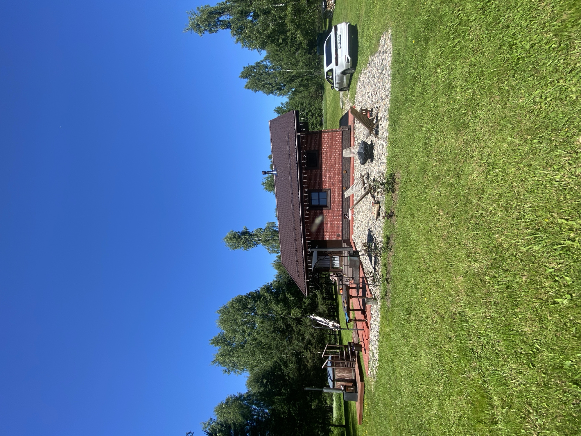 The image captures a rural outdoor scene in Копнино, Russia, under a clear, bright blue sky on a sunny day. A single-story building constructed of red brick with a dark brown gabled roof and a metal chimney stands centrally. Dark-framed windows are visible on the building's facade, and string lights adorn the eaves. To the left of the building, a wooden hot tub with integrated steps and an external stove/chimney is situated on a reddish-brown wooden deck. Adjacent to this deck is a patio area paved with red stones, featuring a dark outdoor table and a folded white patio umbrella. To the right of the building, a light-colored gravel area contains three wooden Adirondack-style chairs positioned around a dark, circular fire pit. A small green plant is rooted in the foreground soil near this gravel area. A white sedan, identified by the Russian license plate 'Y722EK 193 RUS', is parked on the green grass to the right of the building. The property is surrounded by an extensive, well-maintained lawn of green grass and is bordered in the mid-ground and background by a dense forest composed of various tree types, including tall birches and conifers. No individuals are present in the scene. A faint, distant object is discernible in the upper-left portion of the sky.