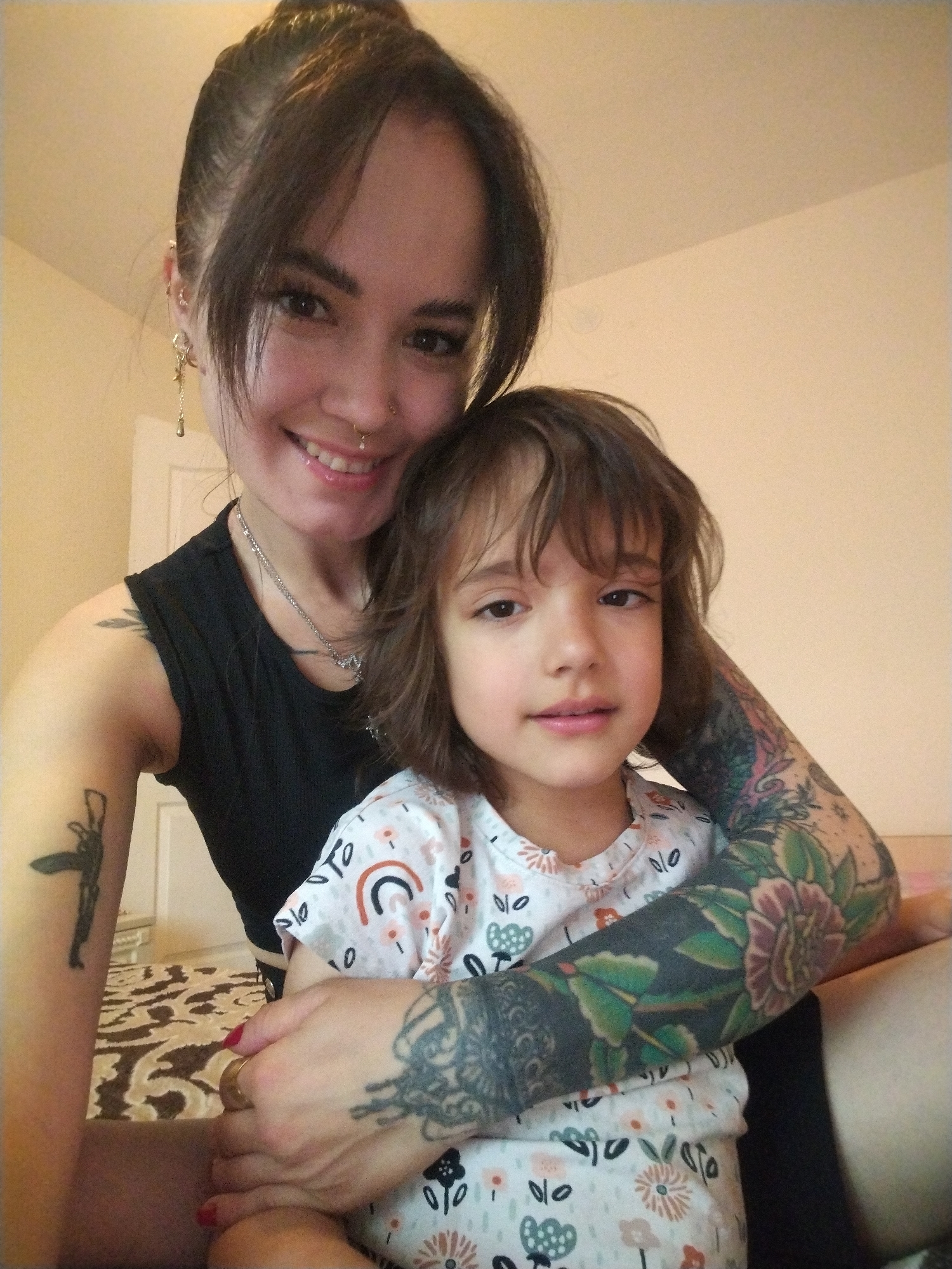 In Noviy, Russia, a woman and a young child are pictured in a close-up, indoor selfie. The woman, on the left, has her hair pulled up and is wearing a black sleeveless top and a silver necklace. She has several visible tattoos on her arms, including a gun on her left arm and floral designs on her right arm. She is smiling and appears to be holding the child. The child, on the right, has brown hair and is wearing a white shirt with a floral and rainbow pattern. The child is looking towards the camera. The background is an indoor setting with a light-colored wall and a patterned rug visible on the floor.