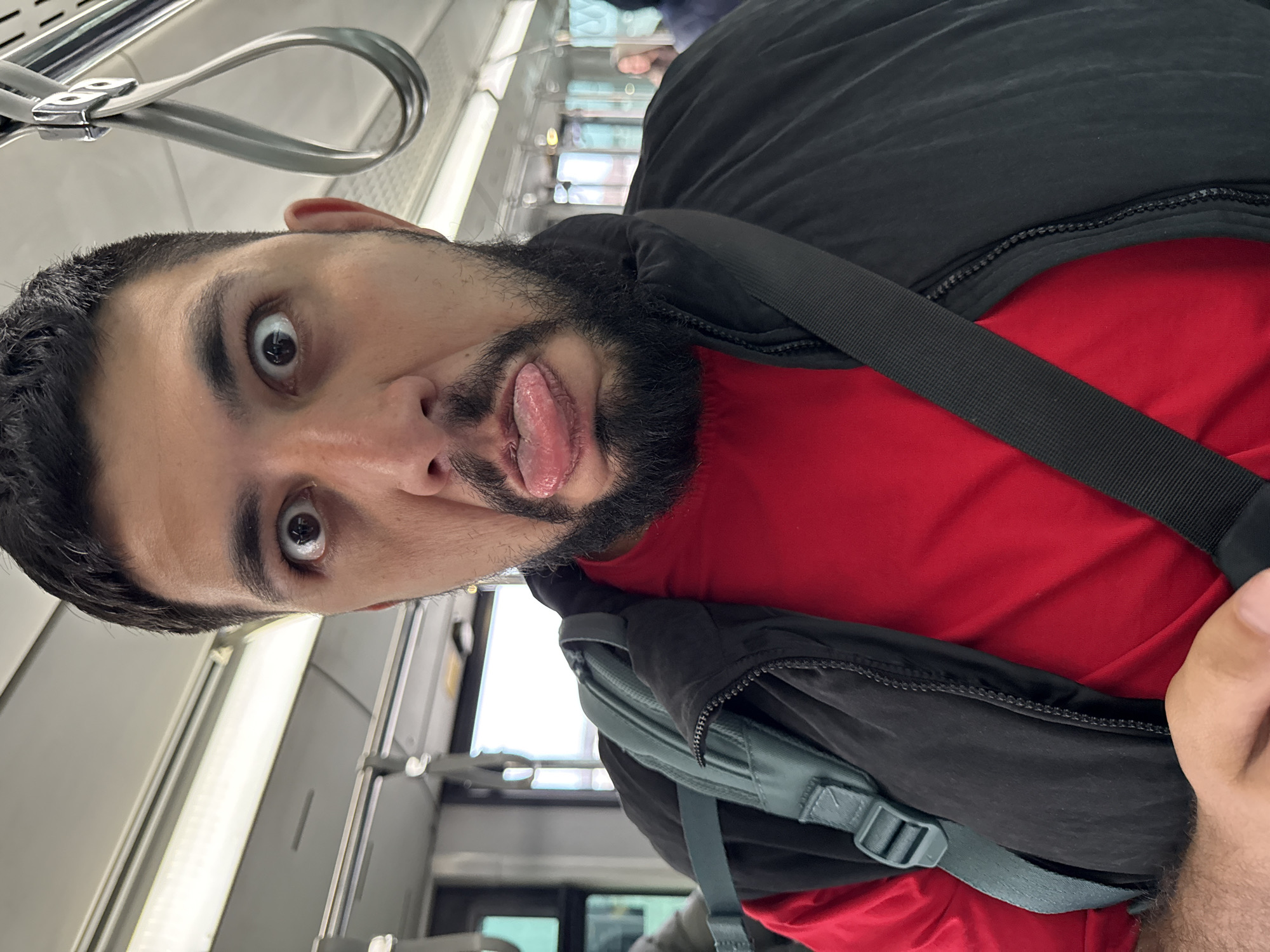 A man with dark hair and a beard is pulling a silly face with his tongue sticking out and eyes wide open. He is wearing a red shirt and a black jacket with a grey backpack slung over his shoulder. The background suggests he is inside a train or bus with rows of windows and overhead hand grips visible. The image was taken from a close-up, slightly tilted perspective. The location in the city of Емельяново (аэропорт), Russia, cannot be determined from the image alone.