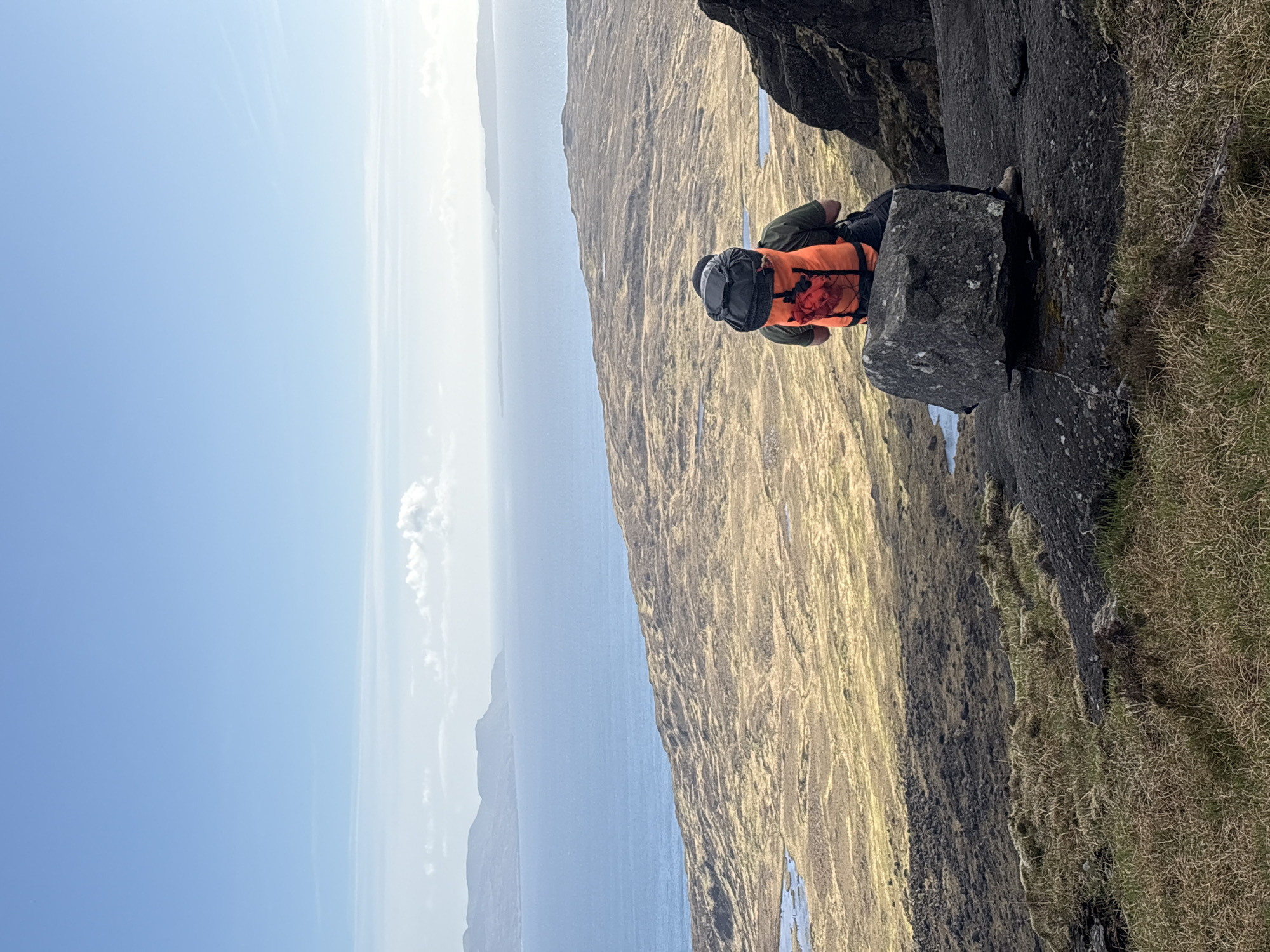 A hiker is resting on a rock ledge on the Isle of Skye, United Kingdom. The hiker wears an orange vest, green pants and carries a grey backpack. The hiker is positioned on a grey rock face. To the left, there is a view of water and distant land under a blue sky. To the right, the rock face is covered in dry grass.