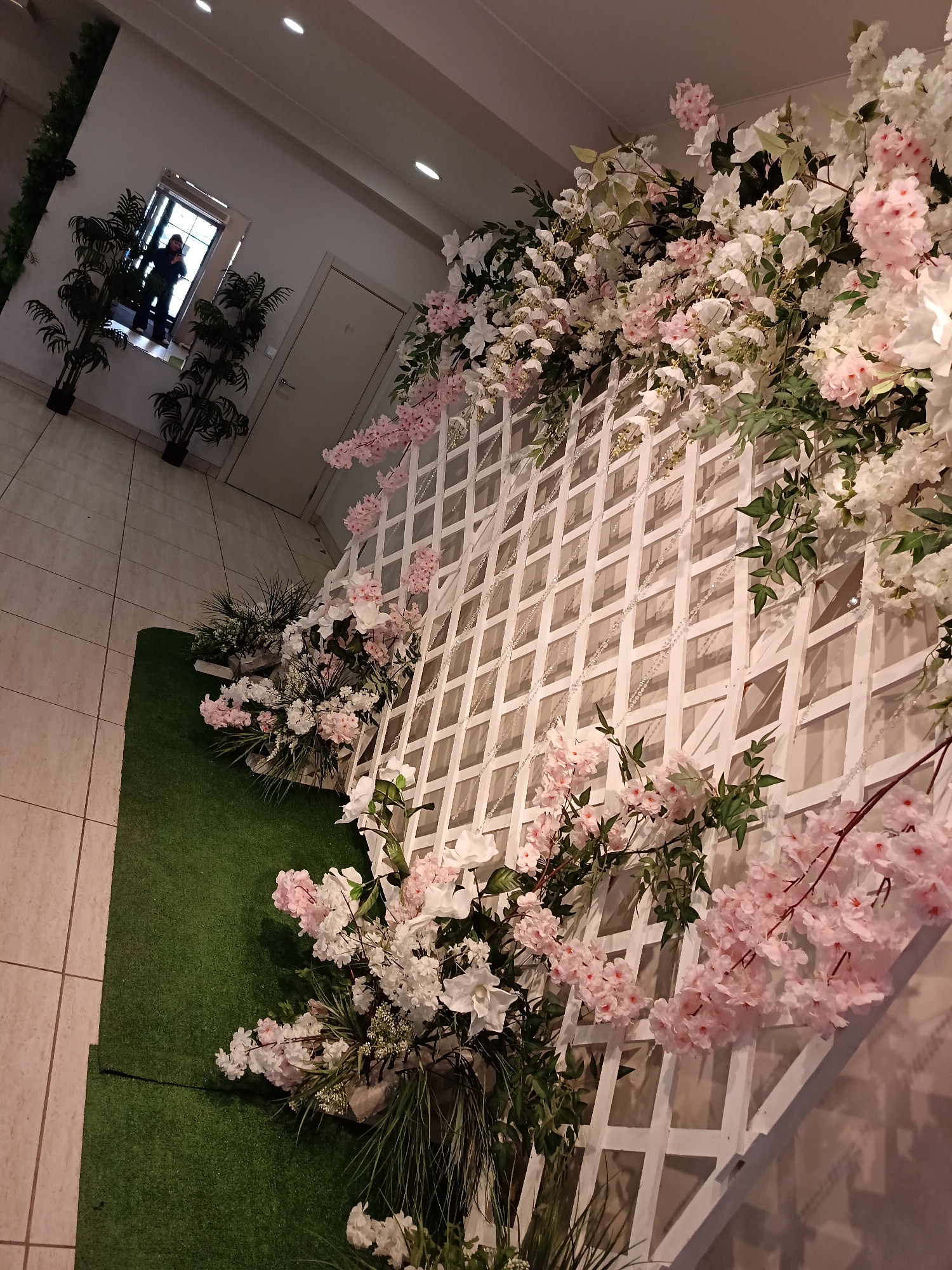 A room with a white lattice wall covered in pink and white flowers and green foliage, with a section of artificial green grass on the floor. In the background, there are potted plants on either side of a window or doorway where a person is visible. The location is possibly Osa, Russia, given the user's request.