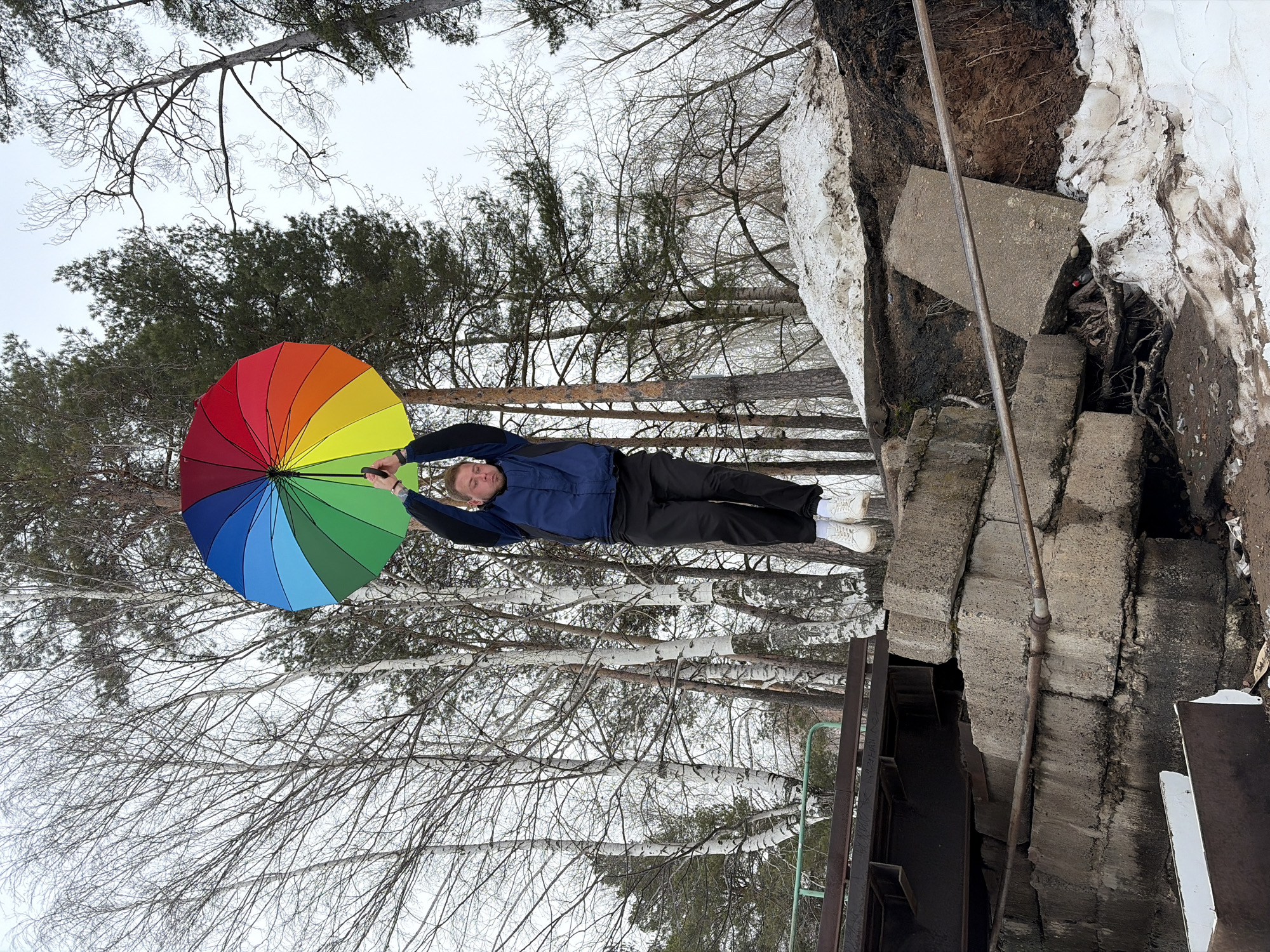 A man in a blue jacket and black pants is holding a rainbow umbrella. He is standing near a series of concrete steps and a snowy embankment in what appears to be a wooded area. The location is Chaikovsky, Russia. The trees are bare, suggesting winter or early spring. The ground is visible between the trees.