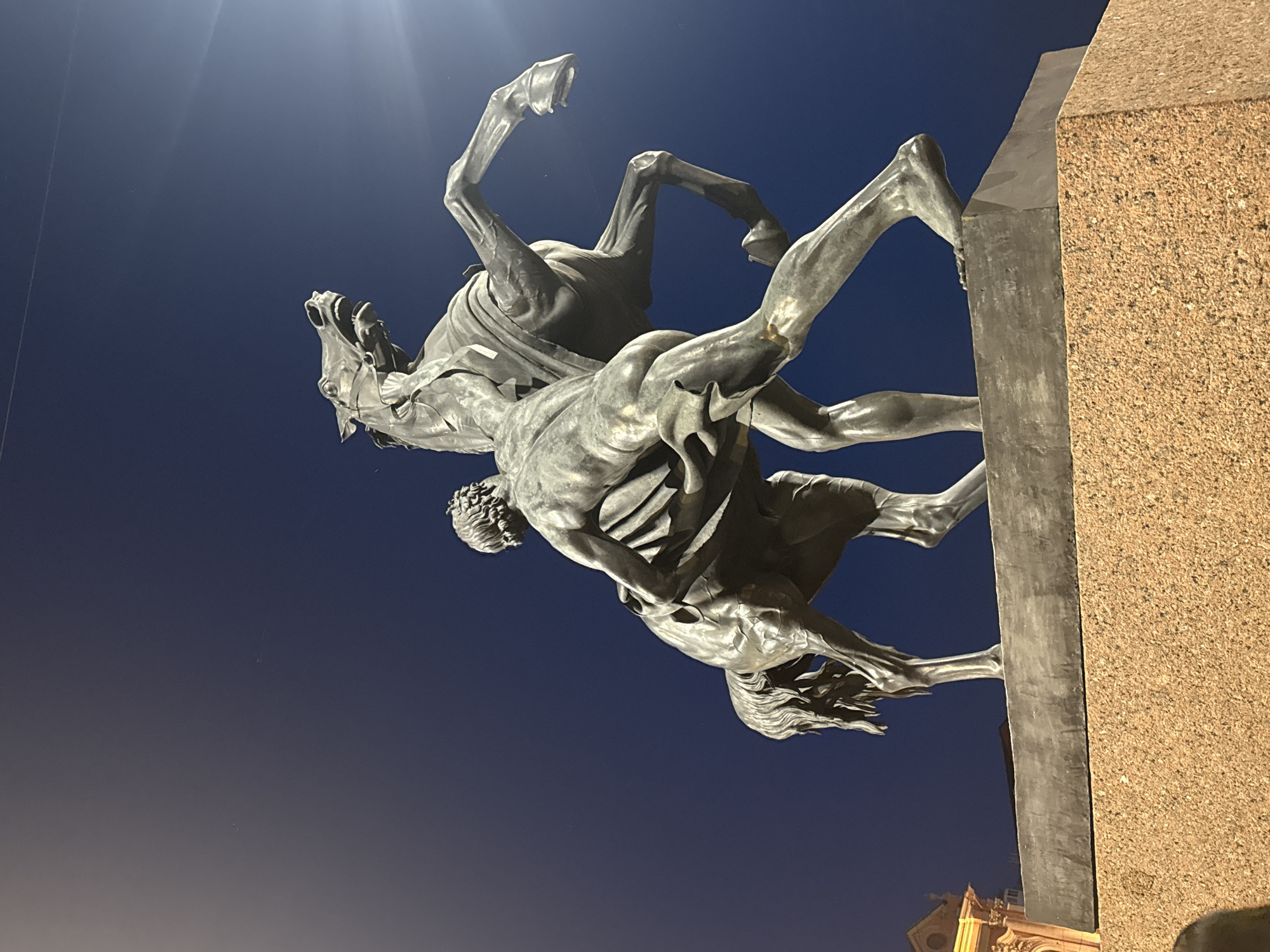 The image shows a bronze sculpture of a man wrestling with a horse, set against a dark blue night sky. The sculpture is located in Saint Petersburg, Russia, specifically at St. Isaac's Square, where it is part of the monument to Nicholas I. The man is depicted with muscular physique, his body contorted as he grapples with the rearing horse. The horse is shown in a dynamic pose, with its front legs raised and its mouth open, appearing to struggle against the man's grip. The sculpture is placed on a stone pedestal, which is illuminated from below, casting subtle shadows. In the background, a portion of a building with neoclassical architecture is visible on the left, its facade lit by warm light. A bright light source is visible in the upper right corner of the frame, creating a lens flare effect. The sky is deep blue, indicative of nighttime, with a few faint streaks, possibly from an airplane or atmospheric phenomena. The overall impression is one of power and struggle captured in a static form.