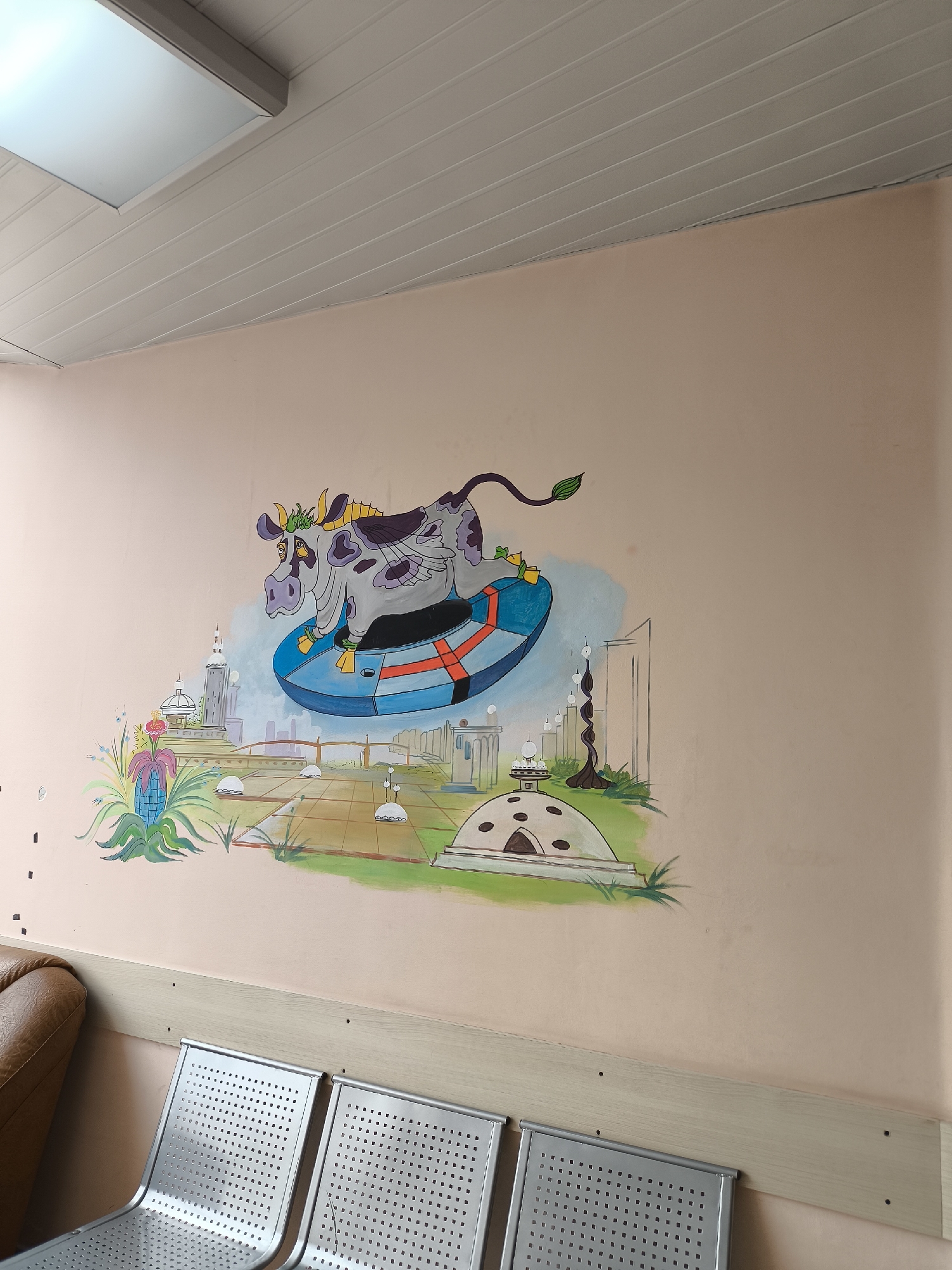 A full shot captures a room with a mural depicting a cow in a UFO floating above a cityscape on a peach-colored wall. A row of silver, perforated metal chairs occupies the foreground, with a tan leather couch partially visible on the left. The mural features a gray and white cow with purple spots riding in a blue and red UFO above a whimsical cityscape with buildings, plants, and structures. The room is lit by a rectangular ceiling light with a white frame. The wall has a horizontal light-colored strip along the bottom where the chairs are placed. The scene is in Khimki, Russia.