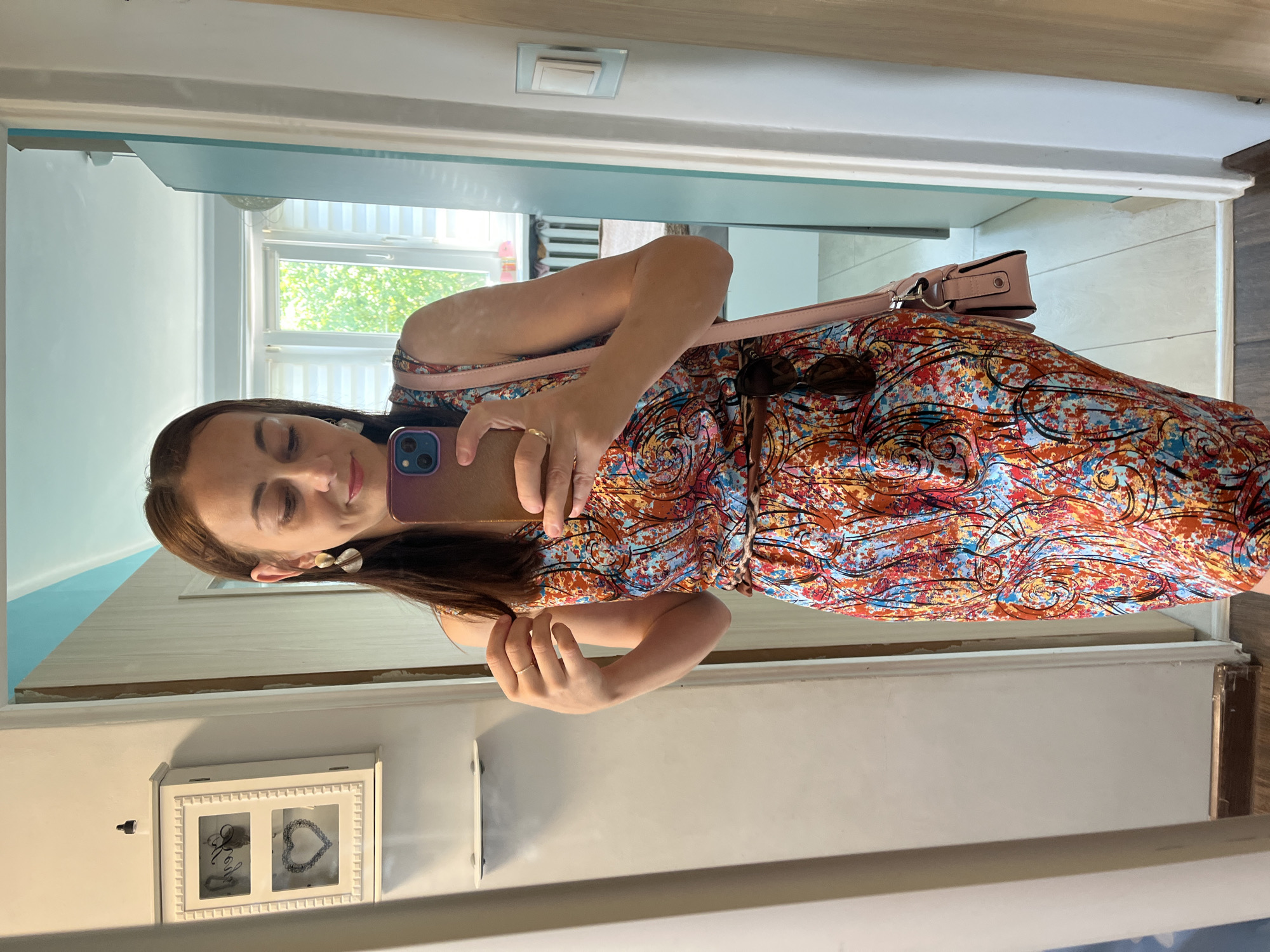 The image shows a young woman taking a selfie in a mirror. She is wearing a colorful, patterned dress and a light pink purse is slung over her shoulder. She has long, dark hair and is wearing a gold, circular earring in her left ear. She is also wearing a thin gold ring on her right hand. In the background, a window with white blinds is visible, suggesting it might be daytime. To the left of the woman, a white wall with a light blue trim is visible, and further to the left, a glimpse of a doorway. Below the woman, a white wall-mounted cabinet with two glass-fronted compartments is visible. The overall setting appears to be indoors, possibly a home or a dressing area. The woman's eyes are closed, and she has a slight smile, suggesting a casual and relaxed mood.