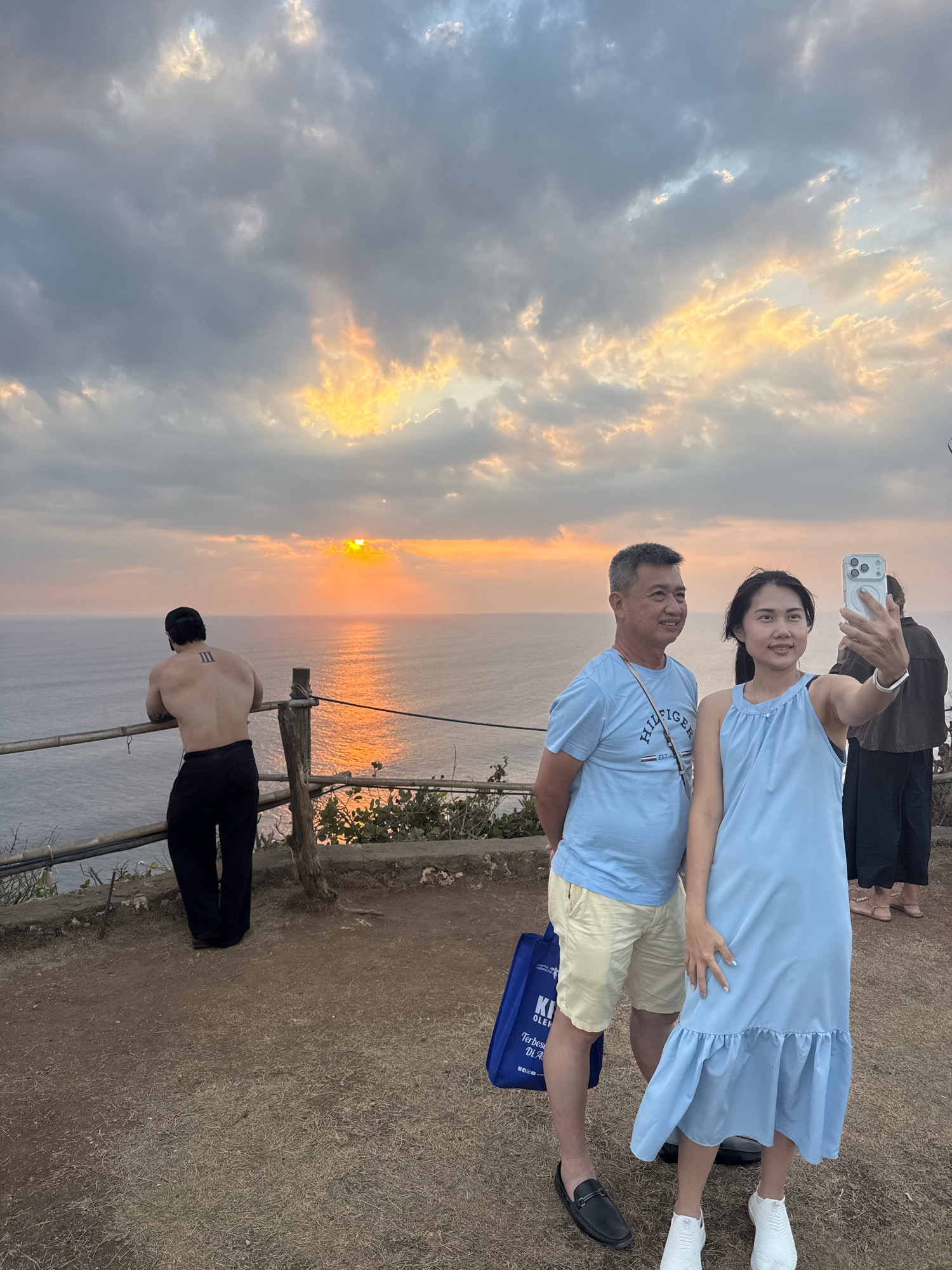 A man and two women are standing on a clifftop overlooking the ocean at sunset. The man in the foreground is wearing a light blue t-shirt and yellow shorts, and he is holding a smartphone up to take a selfie with the woman next to him. The woman is wearing a light blue dress and white sneakers. Behind them, a muscular man with a tattoo on his back is leaning on a railing, looking out at the ocean. The sun is setting over the water, casting a warm glow on the clouds. The ground is covered in dry grass and dirt. There is a wooden railing in the foreground, and a large body of water in the background.