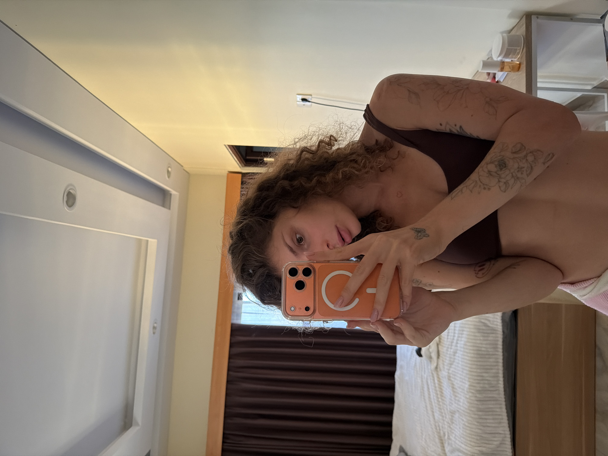 A woman with curly brown hair is taking a selfie in what appears to be a bedroom. She is holding an orange phone with a MagSafe ring on the back. She's wearing a brown top and has tattoos on her arms. The room has a bed with white striped bedding, brown curtains, and a white dresser in the background. The location is identified as North Kuta, Indonesia. The image shows an interior, seemingly private space, with a person documenting themselves via mobile phone.