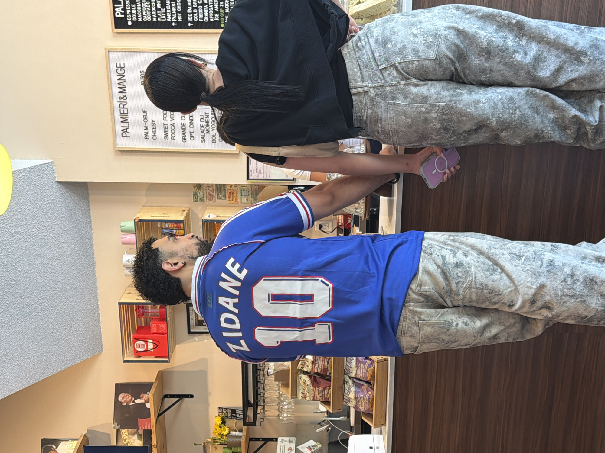 The image shows two people standing indoors in what appears to be a deli or small restaurant. The person on the left is wearing a blue soccer jersey with "ZIDANE" and the number "10" on the back. They have dark, curly hair and a beard. The person on the right has long, dark hair in a ponytail and is wearing a black top and grey jeans. Both are wearing the same grey jeans. The establishment has wooden shelves displaying various food products. A menu board lists items such as "PALMIERI & MANGE" and "PALM-OEUF." The OCR text reveals the name of the Montreal deli as "Palmiéri & Mange."