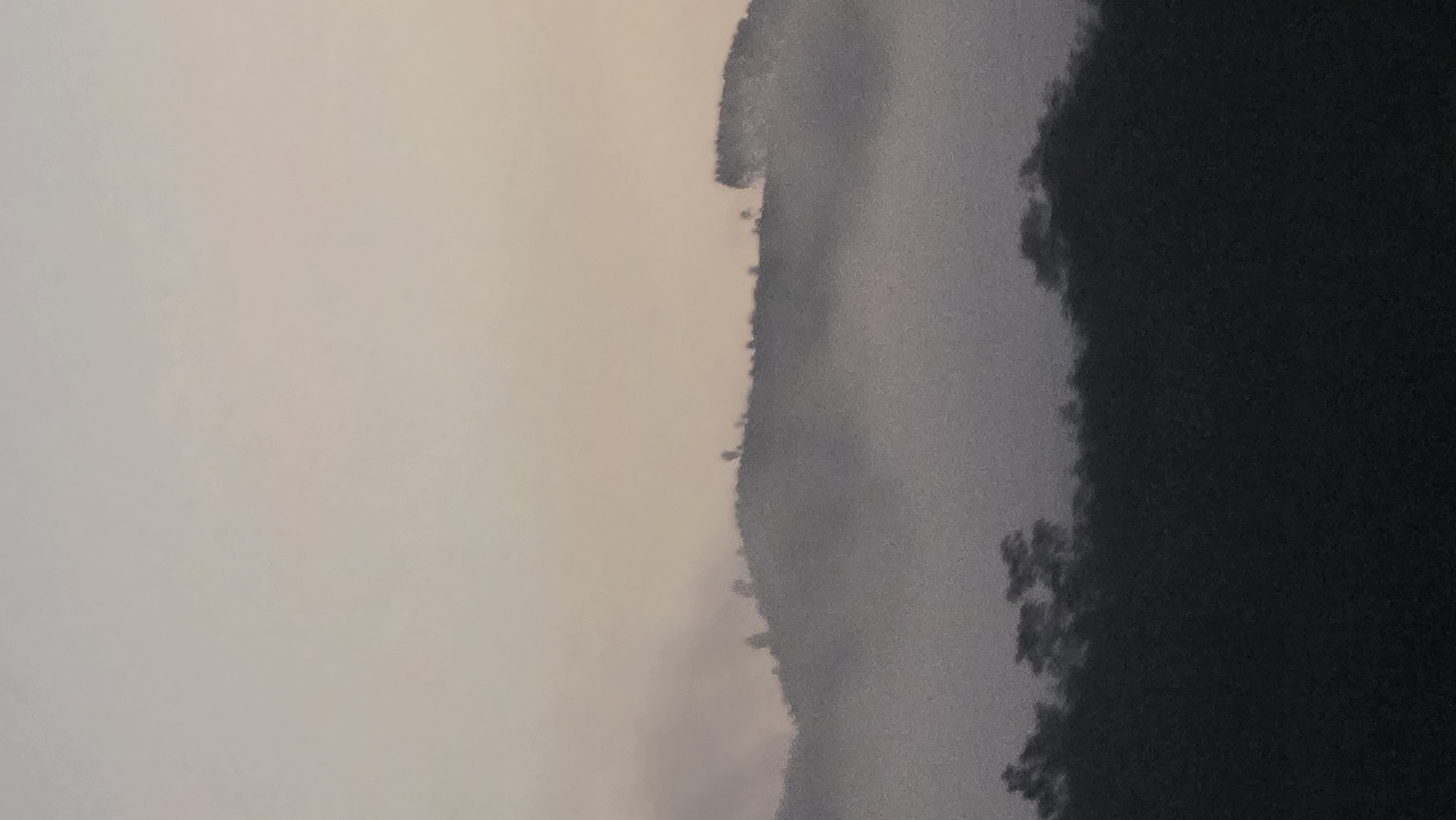 The image depicts a hazy scene in Dak To, Vietnam. On the left side, a light grey or beige sky is visible. Toward the center of the image, there is a dark, irregular shape suggesting a hill or mountain range. The right side of the image is dominated by a very dark area, possibly dense forest or shadow. Details are obscured due to the haziness. There are no people visible. The environment seems remote or natural, given the lack of built structures and the presence of mountainous terrain.