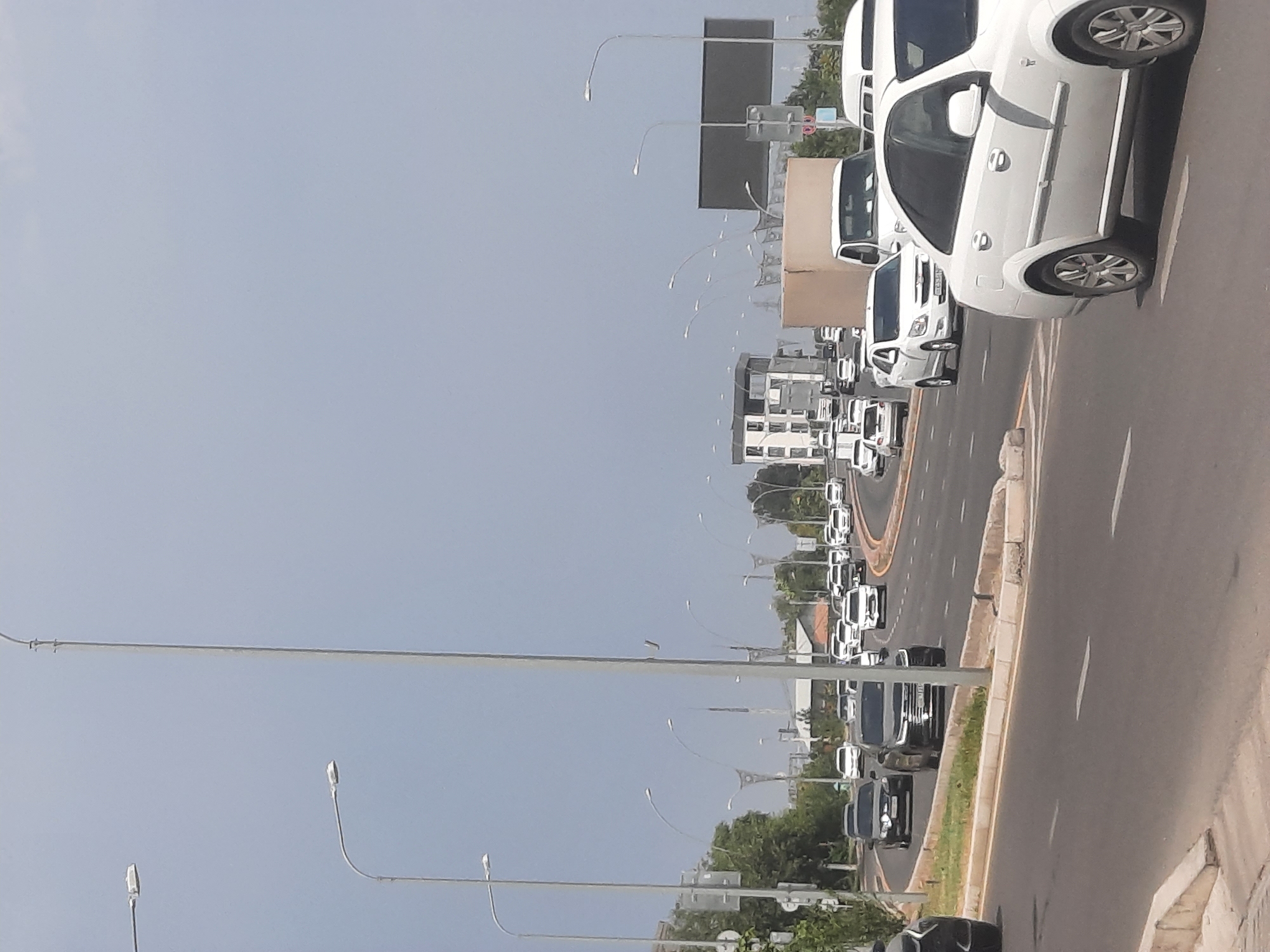 This is a street-level, medium-angle, eye-level shot of a multi-lane road in Tashkent, Uzbekistan, on a clear day. The road curves gently to the right and is filled with traffic. In the foreground, a white sedan is in the right lane. To the left, other cars, including a dark sedan and a dark SUV, are visible. The road is lined with trees and streetlights. In the middle ground, a white truck, a white van, and several other vehicles are driving on the road. In the background, there are buildings and more streetlights. The sky is a pale blue. The scene depicts typical urban traffic in Tashkent.