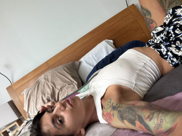 An adult individual is depicted lying on their back on a bed in what appears to be a bedroom in Nusa Dua, Indonesia. The person has dark hair and is looking directly towards the camera. They are wearing a white ribbed tank top or crop top and dark shorts with a white floral or abstract pattern. Extensive, colorful tattoos cover both arms, extending onto other body parts not fully visible. The right arm is bent, with the hand near the head. The left arm is extended downwards relative to the camera's orientation. The bed features a light-colored wooden headboard. Various pillows are present, including a light brown one, a dark blue one, and a white one. The bedding includes a light-colored sheet and a grey or purplish blanket/duvet. The wall behind the bed is light-colored. A black electrical cable is visible on the wall to the left of the headboard. A portion of a light-colored nightstand or small table is partially visible in the lower-left. No other individuals or significant interactions are visible.