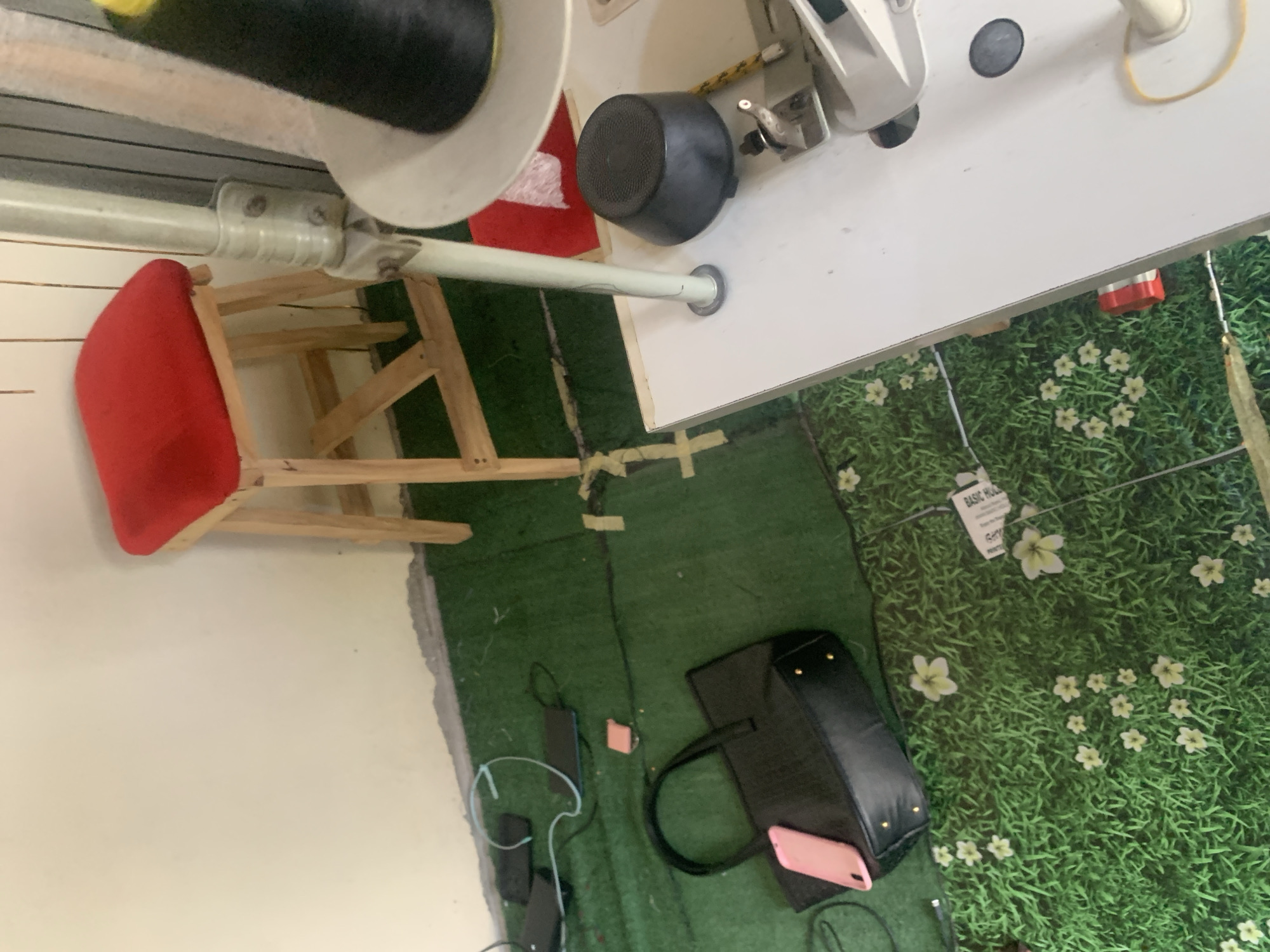 A scene from Jauro Yinu, Nigeria, shows a room with a green carpet resembling grass with scattered white flowers. A red-cushioned stool with a wooden frame sits in the upper right portion of the room. In the foreground, a black leather handbag with a pink phone resting on it is visible on the carpet. Several electronic devices and cables are scattered around the handbag. To the right, part of a white table with a sewing machine is present, featuring a black spool of thread and a black conical object. The walls of the room are cream-colored.