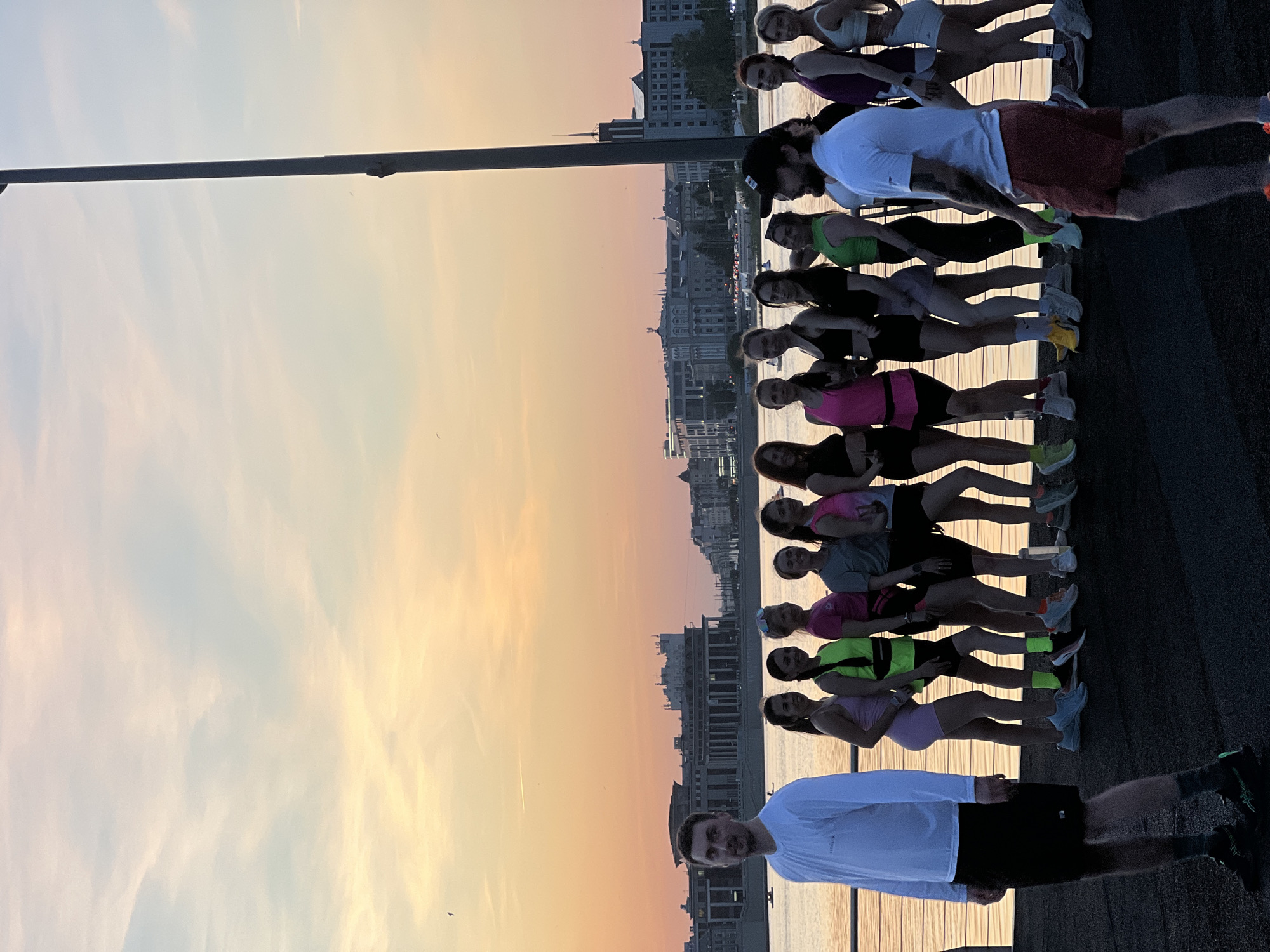 A group of approximately 13 people are posing for a photo on a pier. The pier appears to be made of wood and is located in Kazan, Russia, with city buildings and a body of water in the background. The sky is visible, showing shades of blue and orange. The people are wearing various athletic clothing, including shorts, t-shirts, and athletic shoes.