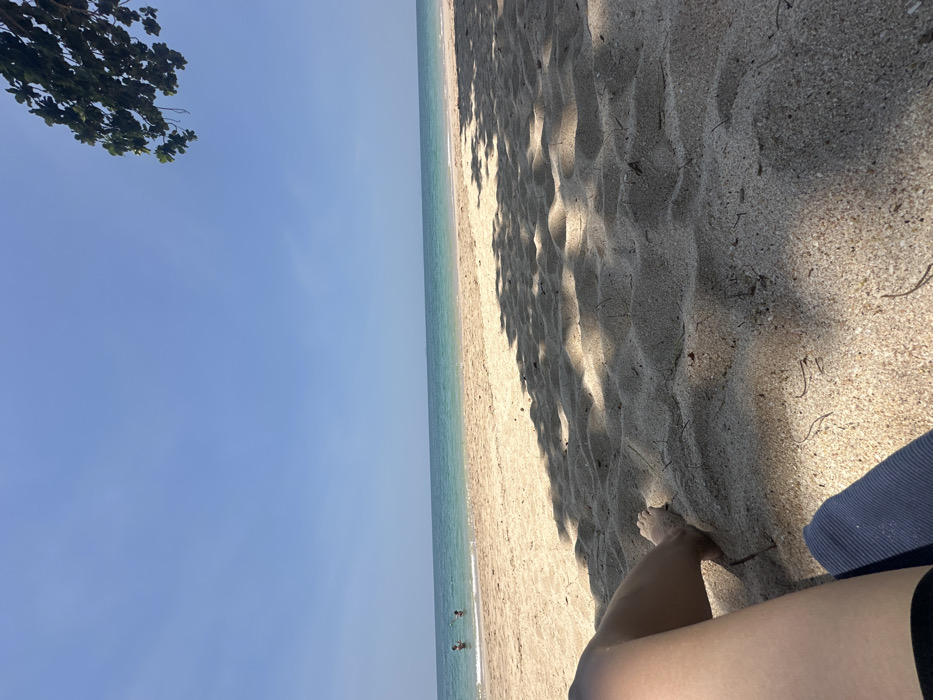 This image depicts a sunny beach scene in Phra Ae, Thailand. The foreground shows the lower legs and bare feet of an individual resting on light-colored sand, with a dark blue, possibly ribbed, fabric item positioned beneath the legs. Dappled shadows from an overhead tree extensively cover the sandy surface, which displays subtle ripples and scattered organic debris. In the midground, the calm, clear turquoise ocean extends to the horizon under a bright blue sky. Two individuals are partially submerged in the water near the shoreline, appearing to be wading or standing. Green foliage from a tree is visible in the upper right corner, contributing to the cast shadows.