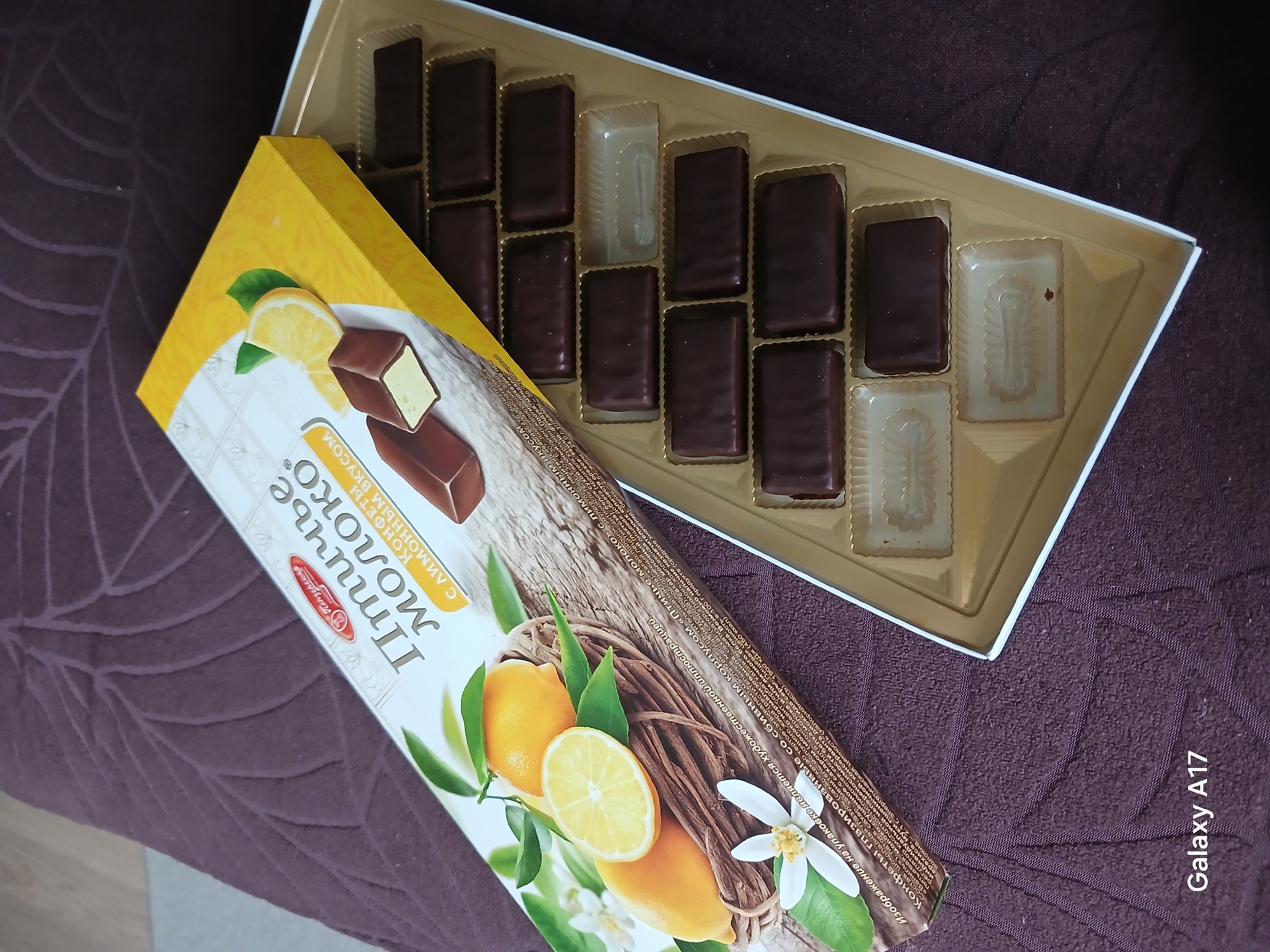 The image shows a box of lemon-flavored "Ptichye Moloko" (bird's milk) candies and its packaging resting on a purple surface with a leaf pattern. Some of the candies have been eaten. The packaging has a picture of lemons and the name of the candy is in Russian. The location of the image is unknown, but the candy is produced by a confectionary based in Penza, Russia. A watermark on the image says "Galaxy A17."