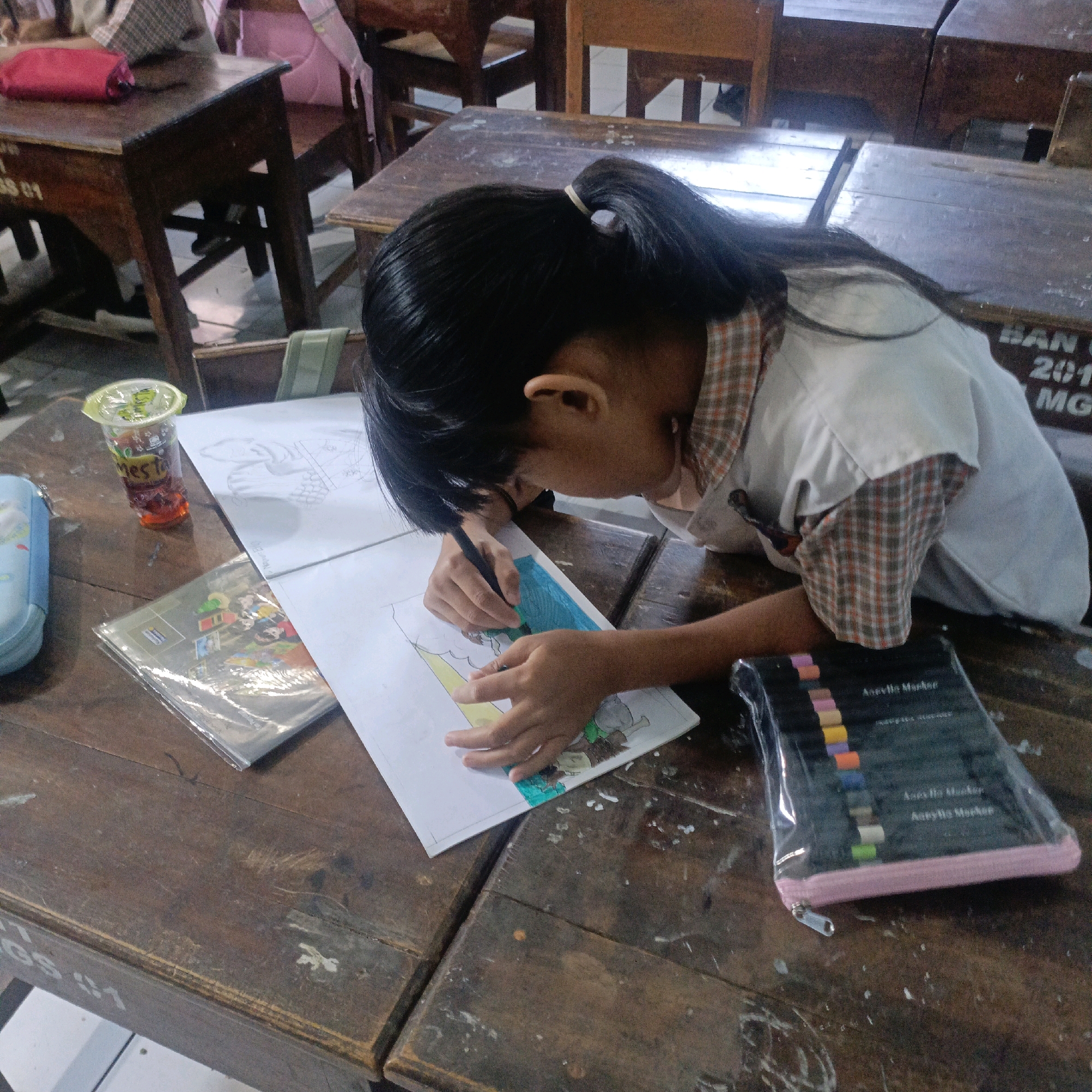 The image depicts a young girl diligently coloring on a piece of paper at a wooden school desk in Salatiga, Indonesia. She is wearing a white school uniform with a plaid collar and cuffs. Her dark hair is pulled back in a ponytail. In front of her, there is a drawing spread out, and she is using a black marker to color a section of it. To her left, there is a clear plastic cup with an orange beverage and a straw, along with a blue pencil case. To her right, a transparent plastic case holds a set of colored markers, arranged in rows. The desk itself is made of dark, weathered wood, showing signs of age and use, with faint white markings on its surface. The background reveals more wooden school desks and chairs, suggesting a classroom setting.