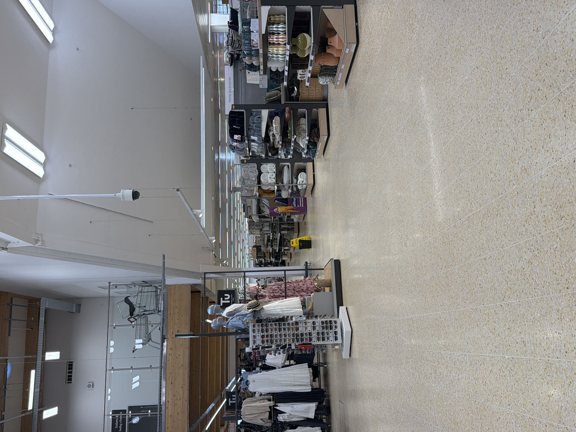 The image shows the interior of a supermarket or department store in the city of Pity Me, United Kingdom. The store has high ceilings and bright lighting from multiple fluorescent fixtures. A security camera is visible. Aisles are lined with shelving units displaying a variety of merchandise, including clothing, accessories, and home goods. Mannequins are dressed in women's clothing. Shopping carts are stacked in the background. The floor is tiled in a speckled pattern. No people are visible in the frame.