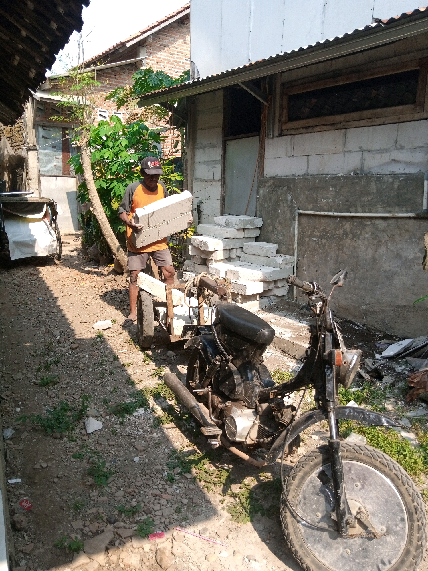 In Batang, Indonesia, a man is seen carrying a stack of concrete blocks. He is dressed in an orange and gray shirt and dark shorts, with a black baseball cap on his head. He stands near a modified motorcycle with a sidecar, which appears to be carrying more concrete blocks. The motorcycle is black with a worn seat and a metal frame extending to support the sidecar. The environment is an outdoor area with a dirt ground covered in gravel and small plants. In the background, there are buildings constructed with concrete blocks and brick, with corrugated metal roofing. A tree with a slender trunk and leafy branches is situated near the man. Another vehicle, possibly a cart or a different type of motorcycle with a sidecar, is parked to the left. The scene suggests a construction or renovation project is underway.