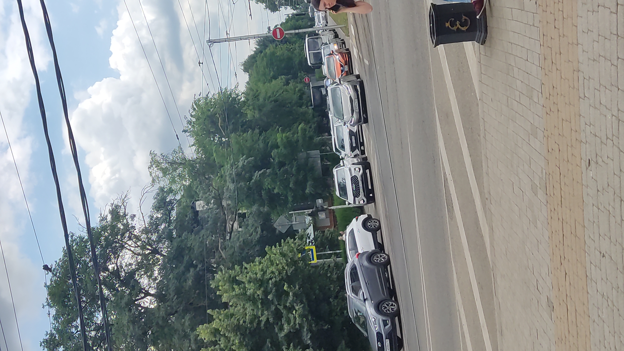 The image captures a street scene in Krasnodar, Russia, with multiple cars in traffic on a multi-lane road. The cars are predominantly SUVs and sedans, with one white SUV in the foreground on the left, followed by a white hatchback. Further back, several other vehicles are visible, including a white SUV with distinctive headlights, an orange sedan, and a white bus. A woman on the right side of the frame is speaking on her phone, and there is a decorative black trash bin on the sidewalk. The environment is urban, with trees lining the background and overhead power lines crisscrossing the sky, which is partially cloudy. The road has clear lane markings and a pedestrian crossing.