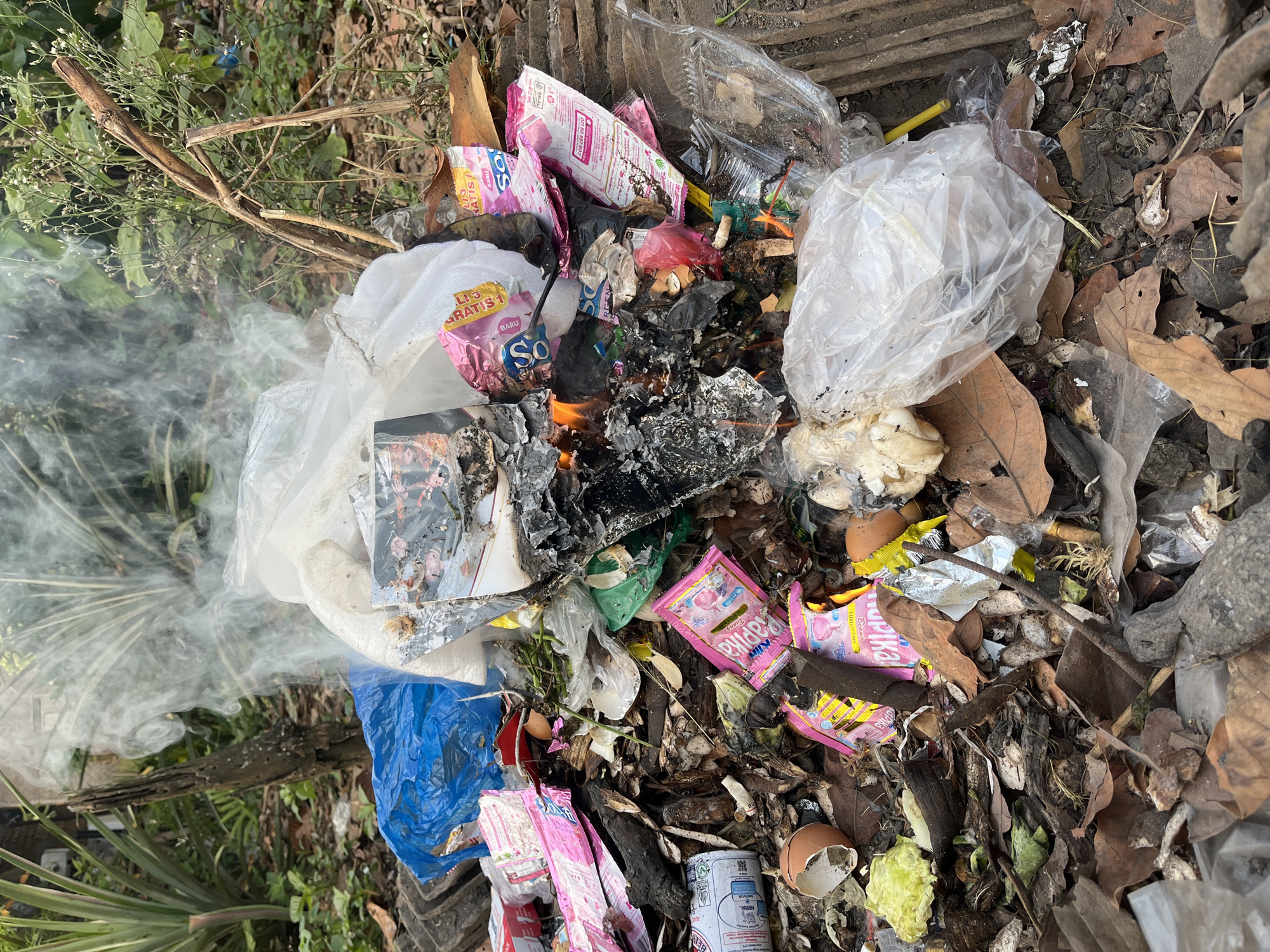 This image depicts an outdoor scene of burning trash, located in a natural or semi-natural environment, possibly on the outskirts or in a less developed area of Kebumen, Indonesia. The trash pile is substantial and composed of various discarded materials, including plastic bags (some blue and transparent), food packaging (such as pink sachets with text that appears to be in Indonesian), burnt paper or cardboard, eggshells, leaves, and other organic debris. A significant amount of smoke is rising from the pile, indicating active burning. The immediate surroundings consist of dry leaves, dirt, and some vegetation, including plants with broad leaves and tall grasses. There are also some dried branches and what appear to be broken pieces of roofing tiles or concrete slabs at the edges of the trash pile. The lighting suggests it is daytime. No people are visible in the image, and the focus is solely on the trash and the fire.