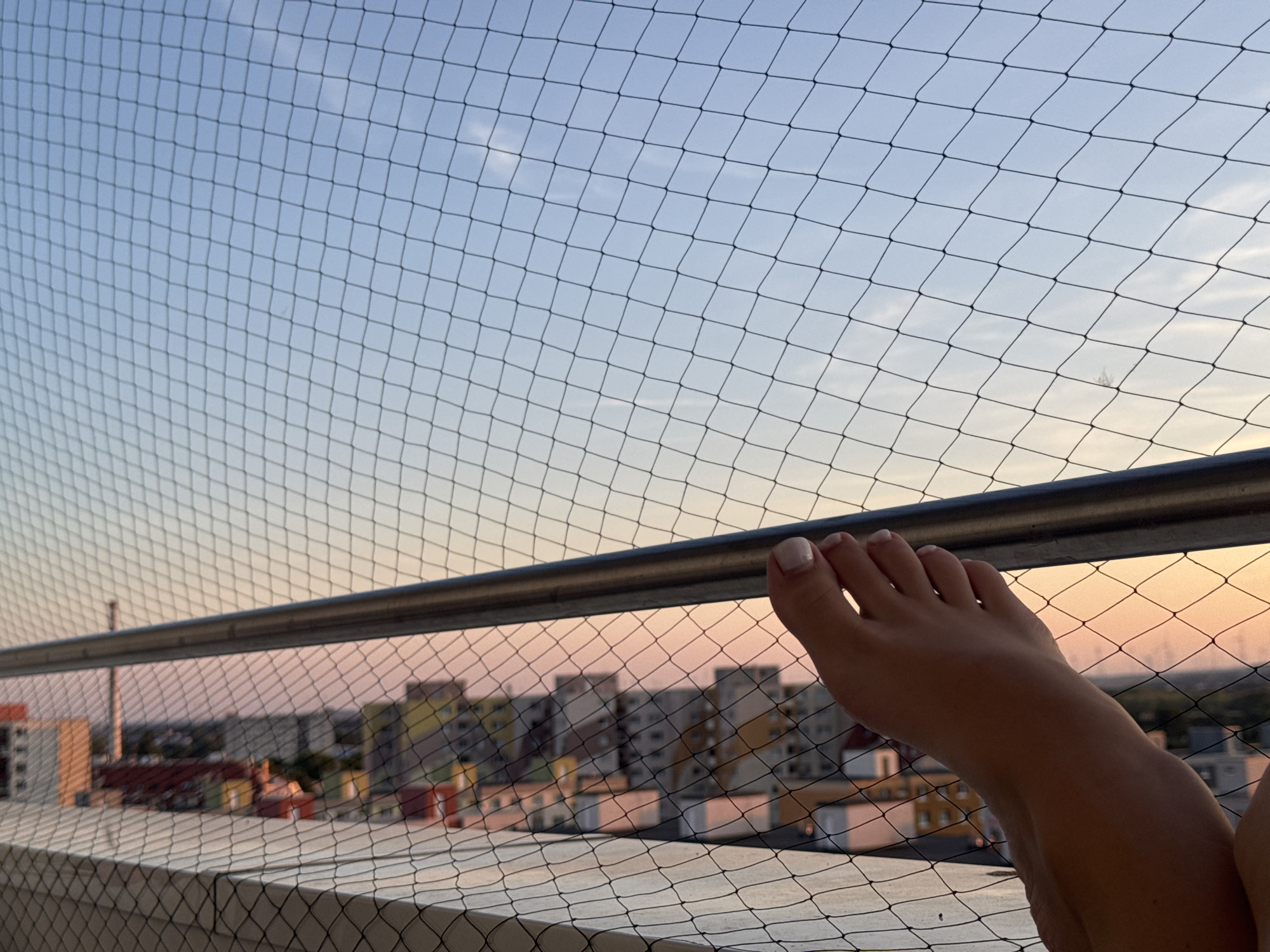 The image shows a close-up of a person's foot resting on a balcony railing, with a net in the foreground and a city skyline in the background. The sky is a soft gradient of blue and pink, indicating either sunrise or sunset. The city appears to be Bratislava, Slovakia, given the architectural style of the buildings. The buildings are of varying heights and colors, some with visible windows and balconies. The overall scene conveys a sense of relaxation and a peaceful evening or morning in the city.