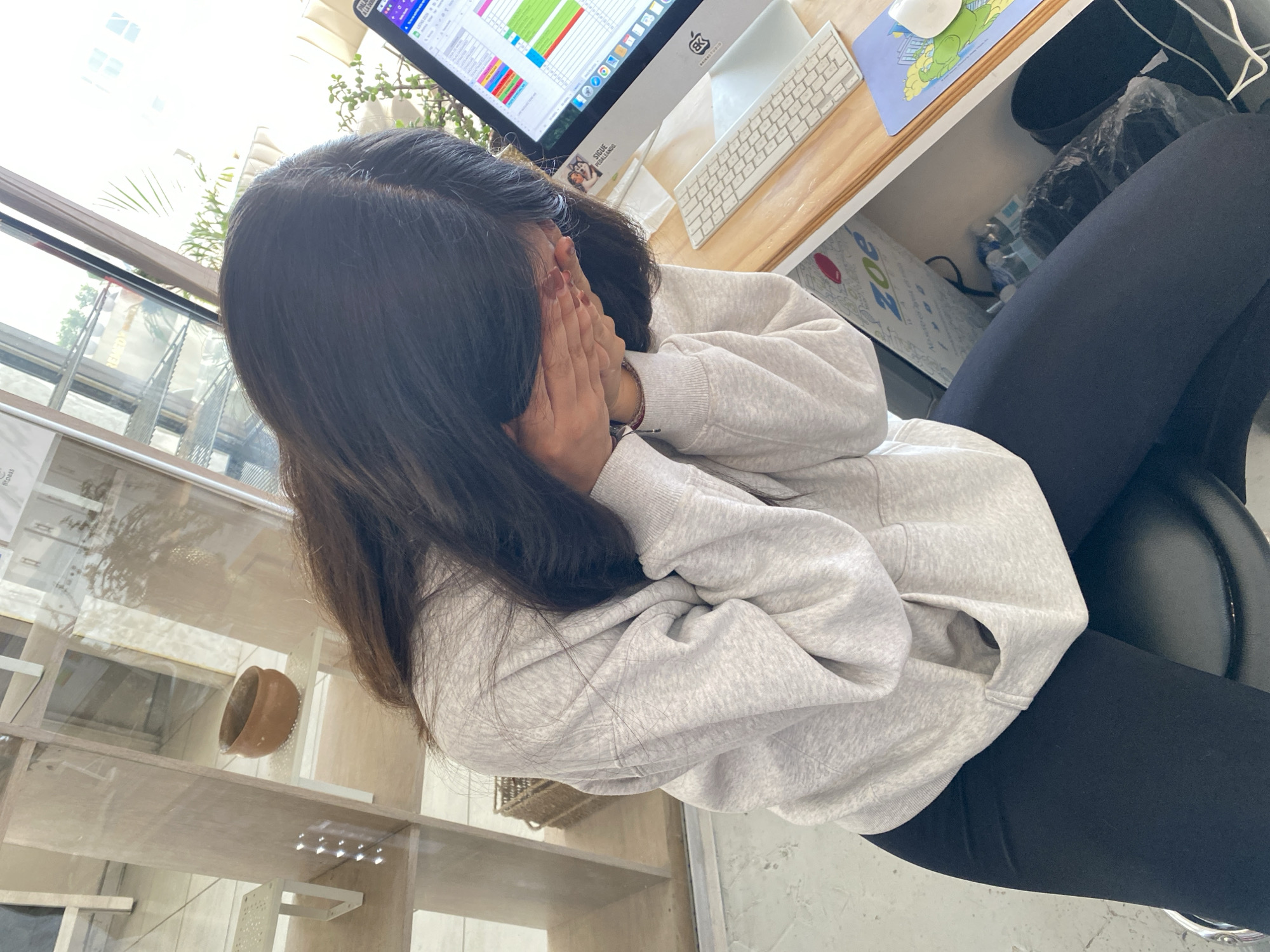 A woman with long, dark hair sits in an office setting in Puebla, Mexico. She is wearing a light grey hoodie and black leggings, sitting on a black office chair. Her face is obscured as she covers it with her hands. She is positioned in front of a desk with a computer monitor displaying data in a spreadsheet, a keyboard, and a mouse on a mousepad. There's also a laptop beside the computer monitor. A shelf unit stands to her left, containing a small terracotta pot. A window is visible in the background.