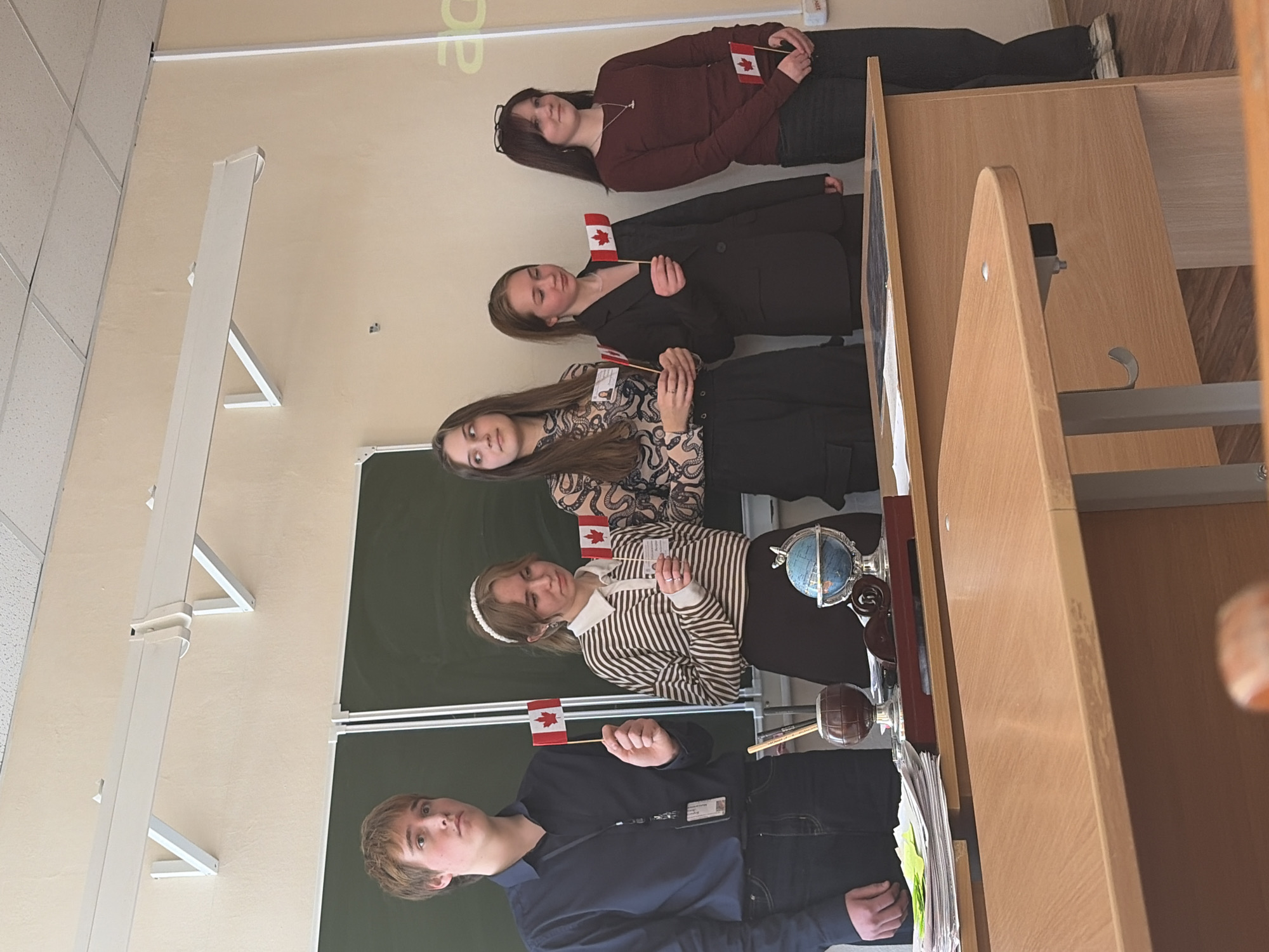 Five individuals, seemingly students, are holding Canadian flags. They are standing in what appears to be a classroom setting, with a chalkboard in the background. The specific location is Chaikovsky, Russia. Objects visible include a globe on a table, papers, and school desks. The individuals are lined up side by side. Their clothing varies, with some wearing casual attire such as sweaters and blouses, while others are in more formal attire, like jackets. There is no explicit action, the scene appears to be static.
