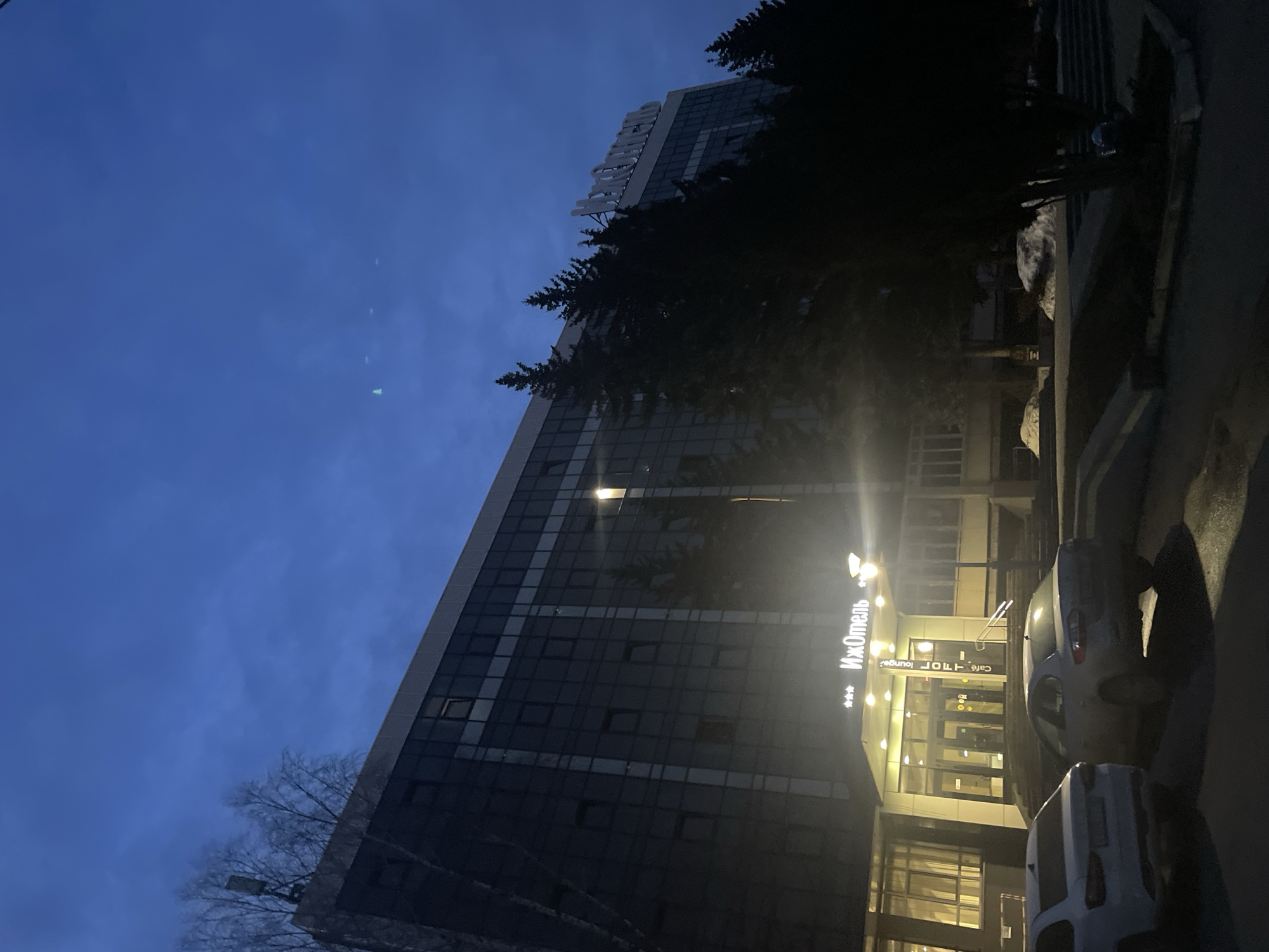 The image captures the exterior of the IzhHotel in Izhevsk, Russia, at dusk or dawn. The building is a modern high-rise with a glass facade, featuring multiple stories of windows. The hotel's illuminated sign is visible near the entrance. A few trees, some deciduous and some evergreen, frame the building. Two cars are parked in front of the hotel. The sky is a deep blue color, indicating that it is close to dawn or dusk. There are no people visible in the image.