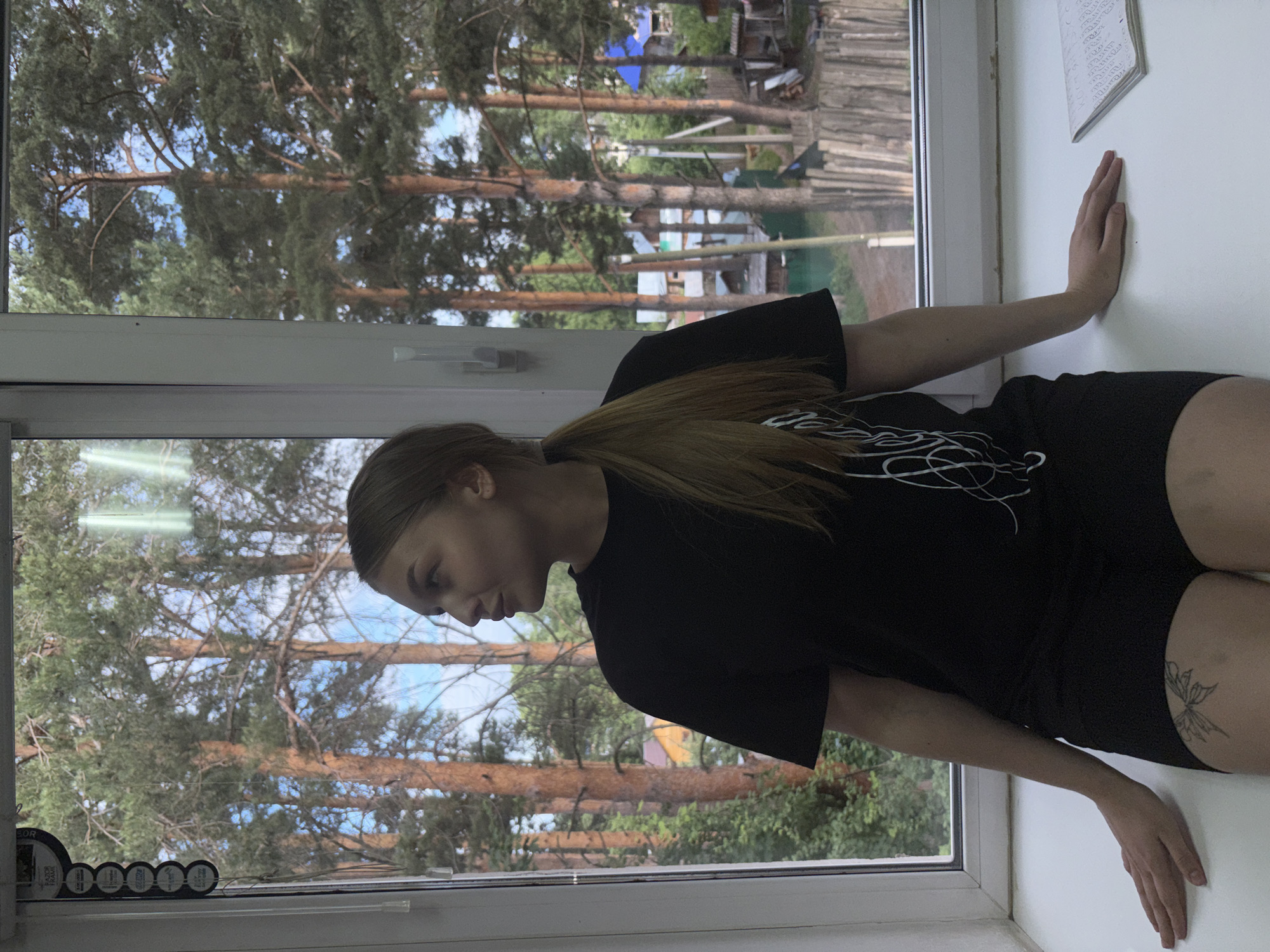 A young woman with blonde hair tied in a ponytail sits on a windowsill, looking out a window at a pine forest and a village. She is wearing a black t-shirt and black shorts. A tattoo of a butterfly is visible on her left thigh. On the windowsill to her right is an open notebook with writing. The setting appears to be a residential area in the city of Chaikovsky, Russia, given the presence of pine trees, wooden fences, and houses typical of the region.