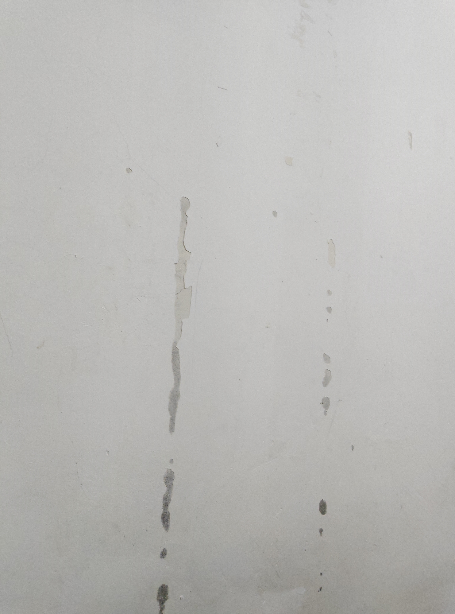 The image shows a close-up view of a pale, textured wall. There are no people or discernible objects in the frame, and no specific scene or setting is depicted. The wall appears to be an interior surface, possibly a wall in a room, but there are no clues to suggest the time of day, weather, or any specific location.
Notable details include several marks and imperfections on the wall. There are streaks and smudges, some of which appear to be water stains or paint drips. Two prominent vertical streaks are visible: one on the left side of the image and another slightly to the right of center. These streaks are irregular in shape and varying in color, with some appearing lighter and others darker gray. There are also smaller, scattered marks and faint lines across the surface of the wall. Some areas show slight peeling or chipping of the surface material. The overall impression is that of a wall that has seen some wear and tear, with evidence of past damage or maintenance issues.
There is no visible text in the image. The lighting is even and diffused, suggesting an indoor environment with ambient light. The focus is entirely on the texture and surface details of the wall.