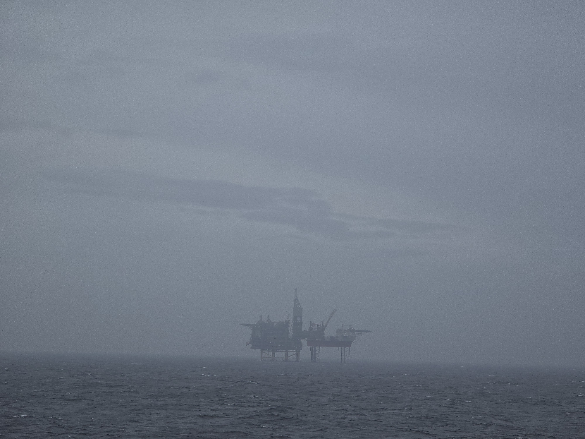 The image depicts an offshore oil platform in a misty or foggy environment. The platform is situated in a body of water with visible waves. The sky is overcast, contributing to the low visibility. There are no visible people or other vessels in the frame. The location is Unknown, Unknown.