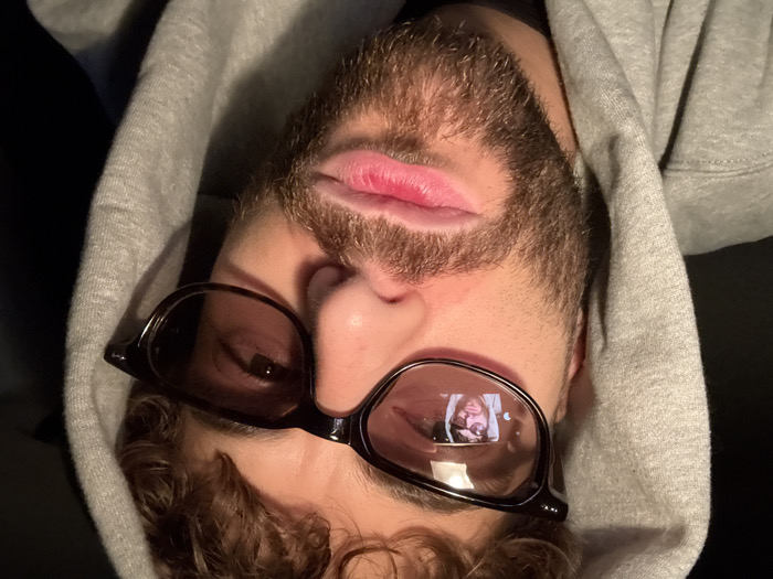 The image displays a close-up, frontal view of a male individual, located in Givors, France. The subject is wearing a grey hooded sweatshirt with the hood pulled up, partially framing his face. He has curly brown hair visible at the forehead, a brown beard, and a mustache. Dark-framed eyeglasses with tinted lenses cover his eyes. Within the left spectacle lens, a distinct reflection shows a smaller, inverted image of a person's face, also wearing a hood and eyeglasses, indicating self-photography. The subject's mouth is slightly parted, with the lower lip appearing full. The background is uniformly dark and featureless, providing no specific environmental details.
