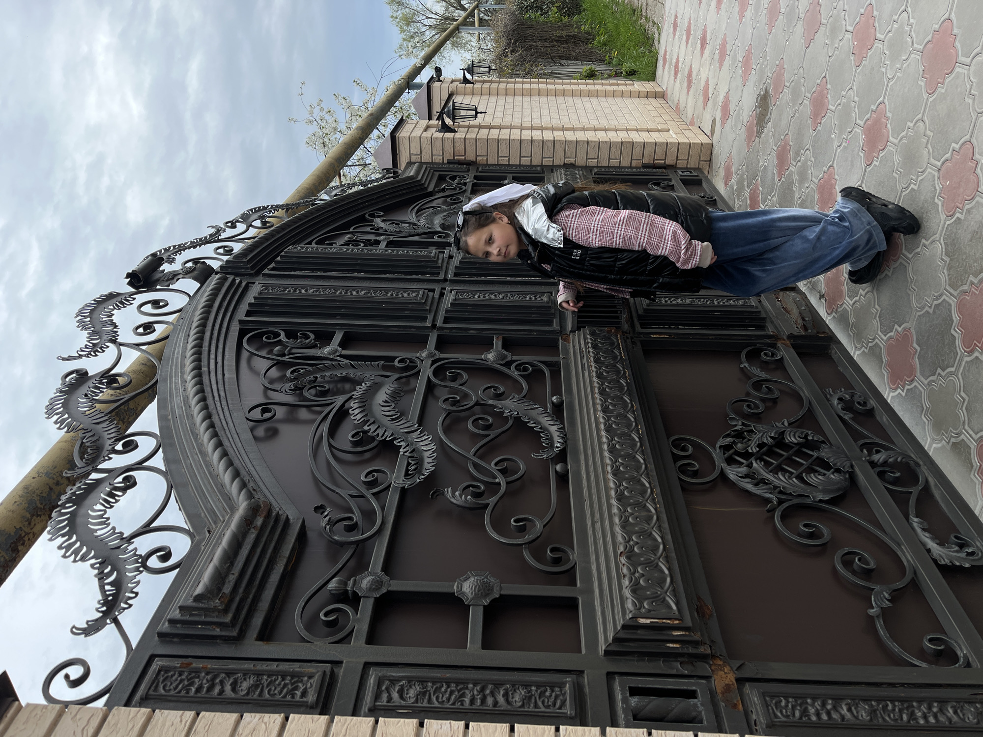 The image, taken in Novopokrovskaya, Russia, depicts a young girl standing beside a large, ornate wrought iron gate, likely marking the entrance to a private property. The image is oriented vertically (landscape view rotated 90 degrees counter-clockwise) but described as if correctly oriented.
The main subject is a girl, appearing to be in her early teens, with dark hair. She is dressed in a white hood pulled up, sunglasses resting on her head, a black puffer vest over a red and white plaid long-sleeved shirt, wide-leg blue pants, and black shoes. She stands with her body slightly turned, looking directly at the camera with a neutral and observant expression. Her right hand rests casually on the gate.
The gate itself is a prominent feature, constructed from dark-colored metal, possibly black or dark brown, with elaborate wrought iron scrollwork, leaf-like motifs, and decorative panels. It shows some signs of weathering and minor rust in places. To the right of the gate stands a sturdy brick pillar adorned with two traditional-style outdoor lamps. The ground in front of the gate is paved with grey tiles featuring a repeating reddish-pink clover-like pattern.
In the background, beyond the gate and pillar, there are trees, some with white blossoms, and green foliage, suggesting spring. A utility pole is visible on the far left. The sky is overcast with soft, light grey clouds, indicating a cloudy daytime. The overall atmosphere is calm, and the girl's attire suggests mild to cool weather. Part of a logo, "AERO", is visible on her black vest.