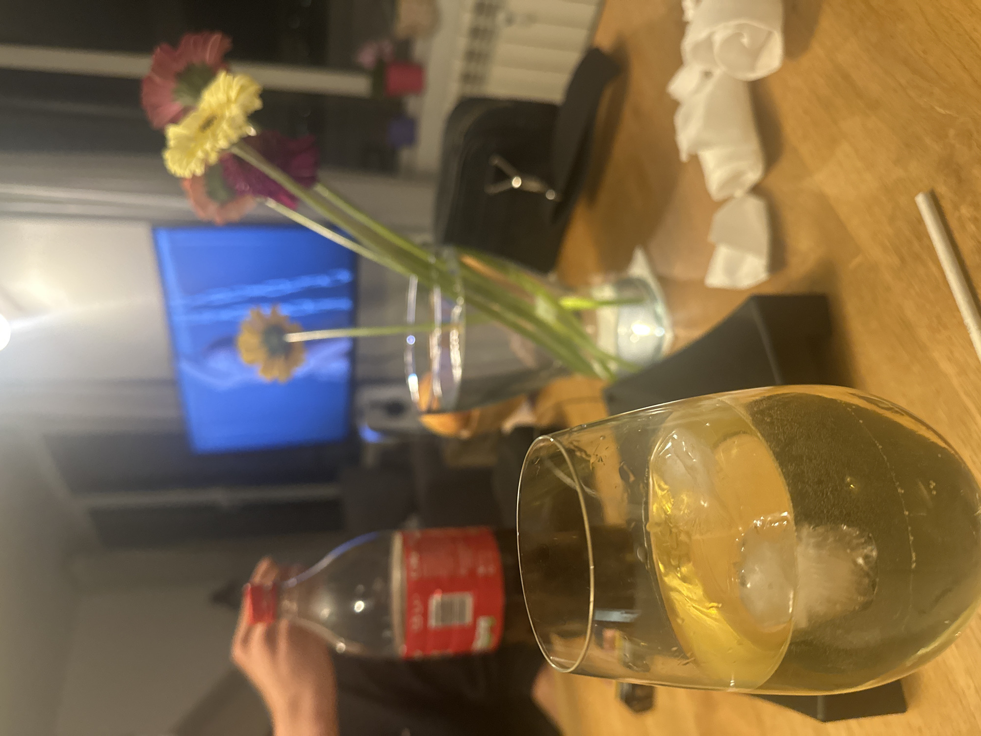 A still life scene is set indoors, likely in Sofia, Bulgaria, given the Cyrillic writing on the Coca-Cola bottle.
The focus is on a glass filled with a light-colored beverage and ice cubes, positioned on a dark, likely rectangular, surface. To the left, a hand is partially visible pouring Coca-Cola from a red and white plastic bottle.
In the background, a vase containing several gerbera daisies of varying colors (red, yellow, and purple) is placed on the table. Behind the vase, a blurred television screen shows an image.
Other objects on the wooden table include a black case, crumpled white napkins, and a white cigarette. The environment suggests a casual, domestic setting.