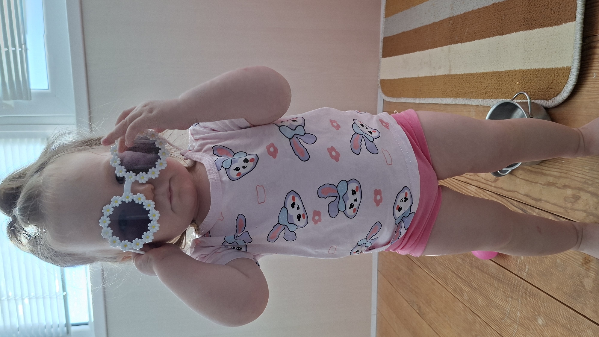 A child wearing pink rabbit-themed clothing and flower-rimmed sunglasses poses in an indoor setting. The location is Chaikovsky, Russia. The child touches the sunglasses. A striped rug is visible in the background. A metal bucket rests on the wooden floor. The child is positioned near a window.
