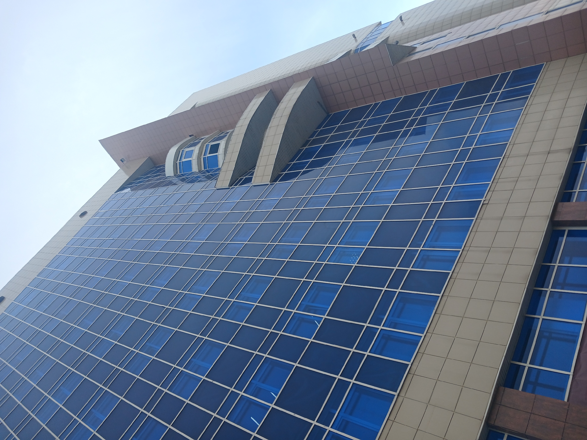 The image captures a low-angle view of a modern, multi-story building in Perm, Russia. The building is characterized by its predominantly blue glass facade, creating a grid-like pattern with the window frames. It features architectural details like protruding elements and curved structures near the top. The sky is overcast, providing diffused lighting to the scene. The building has a contemporary design with a combination of glass and neutral-colored solid panels, adding to its aesthetic appeal.