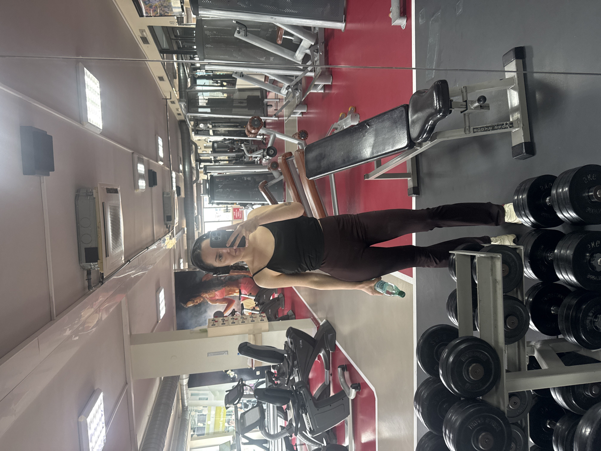 A woman in a black tank top and brown leggings holds a phone in one hand and a water bottle in the other, standing in a gym. The gym contains various exercise machines, including weight racks and benches. The gym's color scheme includes red and grey. There is a mural on one wall. There is no indication of the city of Burgas, Bulgaria.