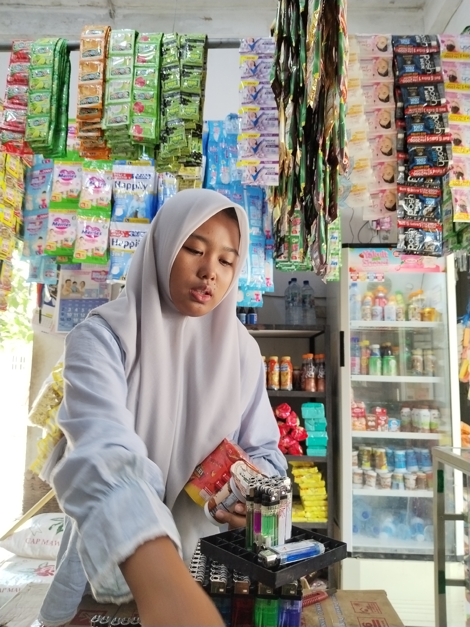 This image captures a young woman wearing a light blue hijab and a matching light blue collared shirt in what appears to be a small convenience store or warung in Sawit Seberang, Indonesia. The woman is the central subject, holding a red packet in her right hand and a lighter display in her left. She is looking down, engaged in an activity not fully discernible from the image, though her posture suggests she might be arranging or examining the lighters.
The background is densely packed with merchandise, typical of such stores. Shelves are stacked with various packaged goods, including what appear to be small sachets of food products, detergents, or personal care items hanging from racks above. To the right, a glass-doored refrigerator displays rows of drinks and other items. The environment is brightly lit, likely from artificial lighting within the store, with some natural light possibly entering from the left. The overall impression is of a busy, well-stocked retail space.