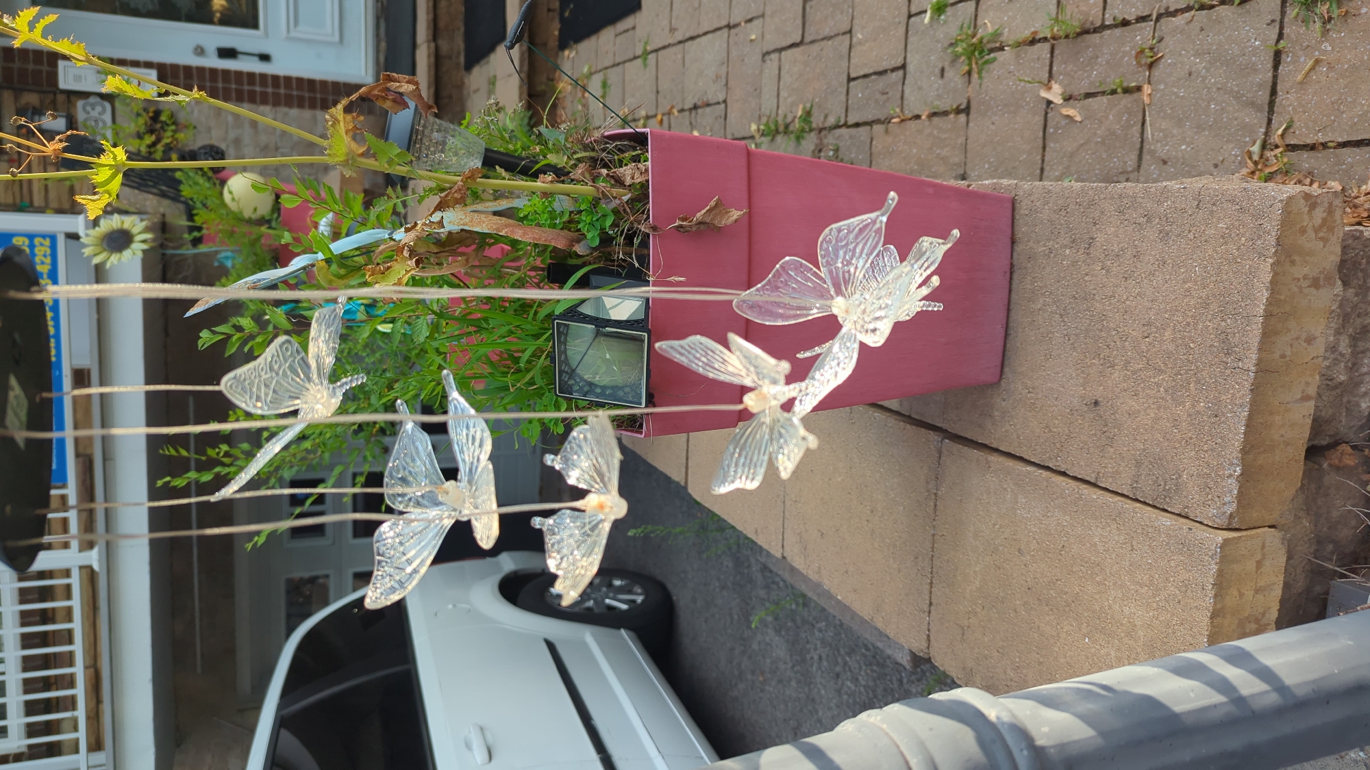 In Montreal, Canada, an outdoor scene depicts a plant pot holding greenery and a solar-powered light. Hanging from above are decorative glass butterflies. A white van is parked in the background, and a brick walkway is visible.