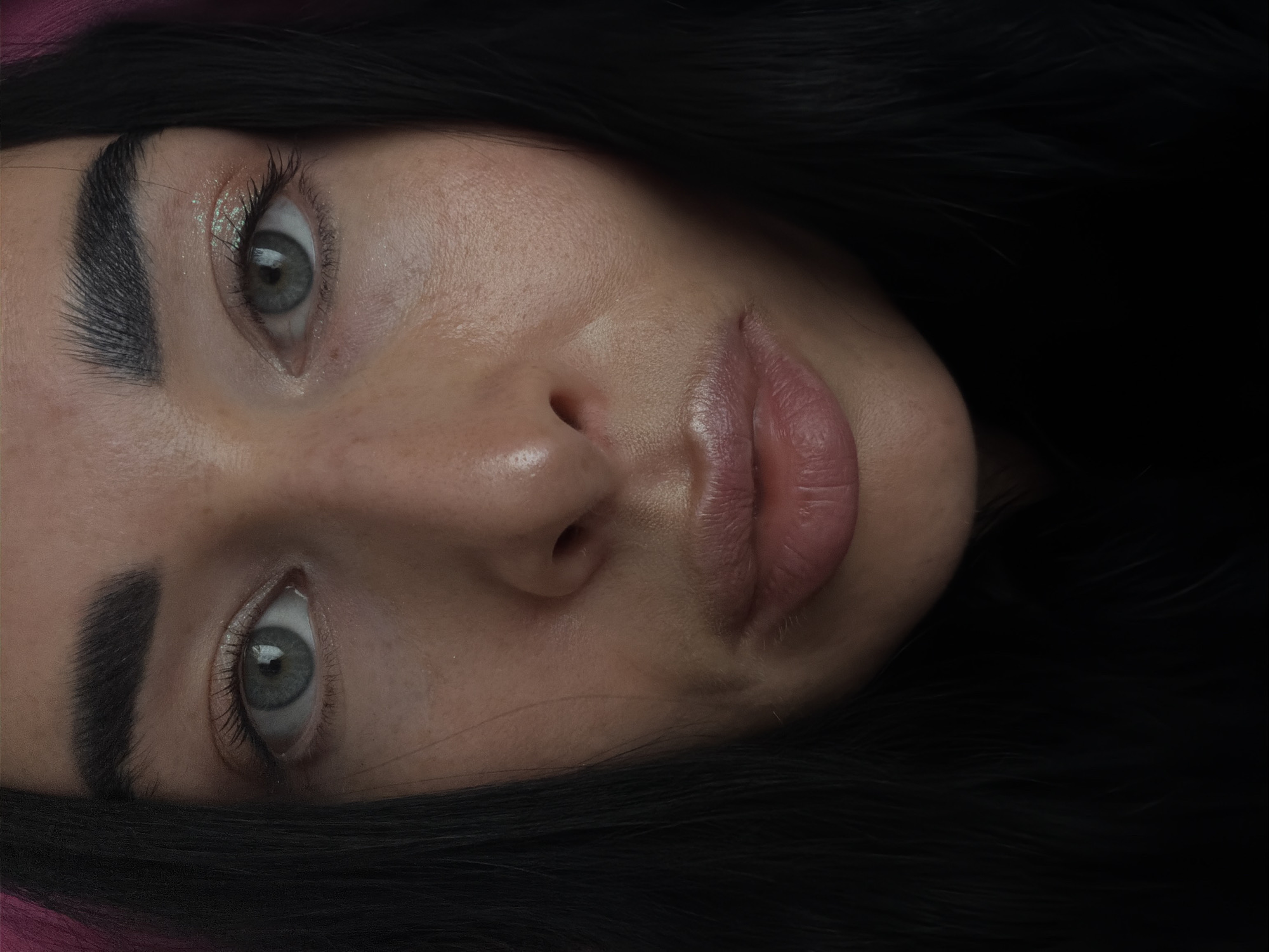 A close-up portrait shows a person's face tilted, with gray eyes and thick eyebrows. Their skin appears to have a dewy sheen, and their lips are full. The person has dark hair that frames their face. The background consists of areas of shadow and a hint of pinkish color near the top. There is no visible indication of the location being Saint Petersburg, Russia.