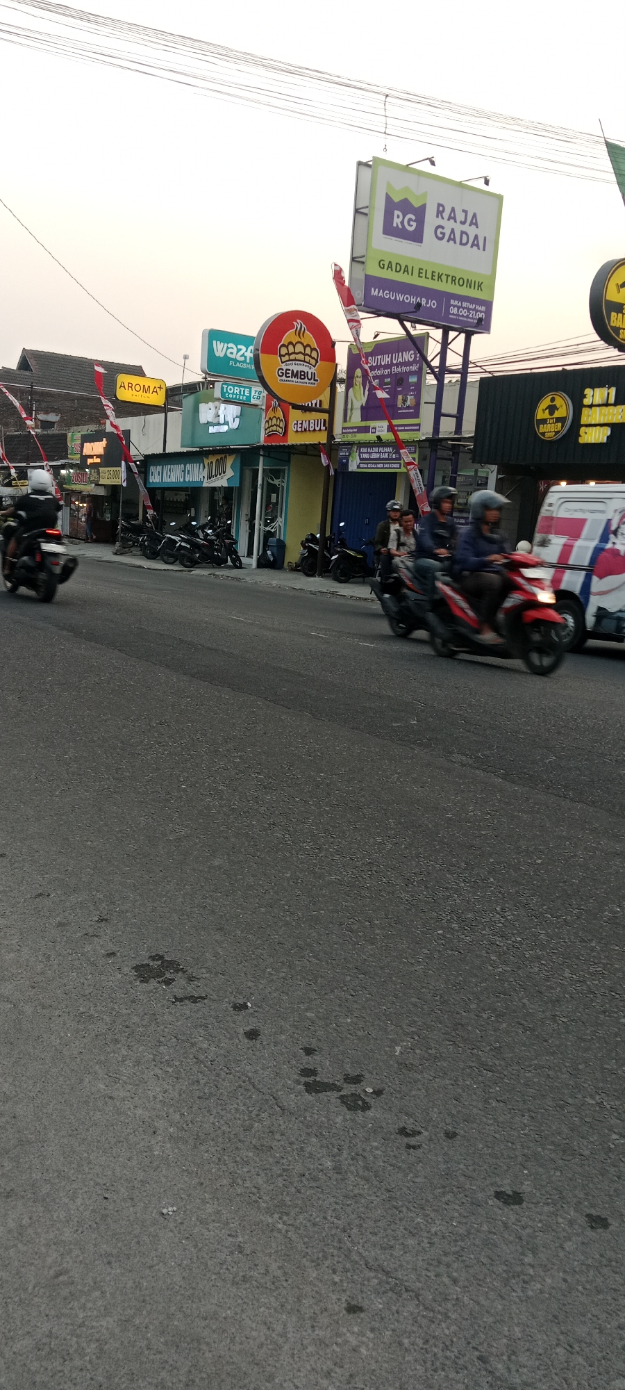 The image depicts a street scene in Maguwoharjo, Indonesia, likely during daylight given the bright sky. The foreground is dominated by an asphalt road with some dark stains. On the right side of the road, a white van with red and blue graphics is visible. Two motorcycles are in motion on the road, with one rider in the foreground on a red motorcycle and another group of riders on motorcycles further back.
Along the left side of the road, several shops and businesses are visible, indicated by various signs. Notable signs include "AROMA+", "WAZ.FLA.SHIP", "GEMBUL", and "TORT COFFEE CO". One storefront has text that reads "CICI KERING CUMA 10.000". In front of these shops, several motorcycles are parked.
A large billboard is prominently displayed above the shops, featuring the text "RAJA GADAI GADAI ELEKTRONIK" with the location "MAGUWOHARJO". Another billboard above it has "RG" in a green box. Further down the street, another business sign reads "3IN1 BARBER SHOP". The background shows a bright, possibly hazy sky with power lines crossing. Red and white striped flags are also visible attached to some of the establishments. The overall impression is that of a busy commercial street.