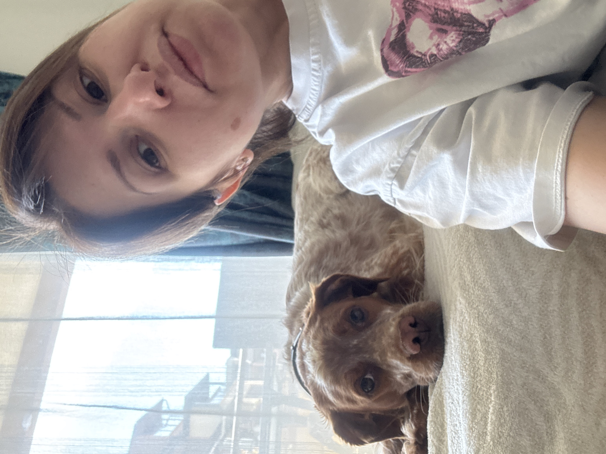 A selfie depicts a woman and a brown dog indoors. The woman is positioned in the upper left, and the dog occupies the lower right. She is wearing a white t-shirt with a pink graphic. The dog is on a light-colored fabric, possibly a sofa. A window is visible behind them, partially obscured by blinds. The location cannot be determined from this image, therefore I can not confirm this image is from Sofia, Bulgaria.