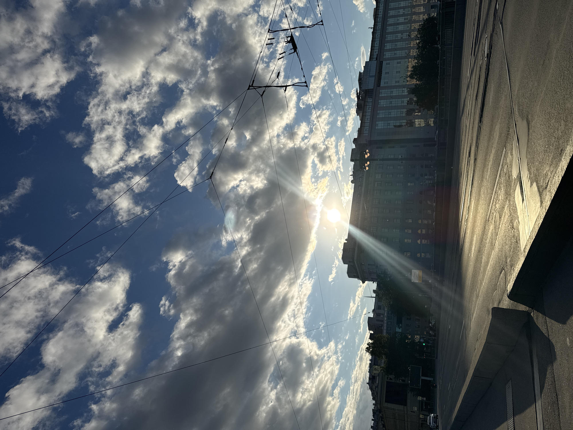 The media file captures a low-angle, street-level view of an urban scene in Saint Petersburg, Russia, on a bright, cloudy day. The dominant elements are the sky, overhead power lines, buildings, and a paved street with tram tracks. The sun is low in the sky, casting a strong glare and creating visible sunbeams that cut through the clouds and illuminate the scene.
The sky is a vibrant blue, filled with a mix of fluffy white and darker grey clouds, suggesting a dynamic weather pattern. Numerous power lines crisscross the sky, supported by visible electrical infrastructure, hinting at the presence of public transportation like trams.
On the right side of the image, a large, multi-story building with a modern facade, characterized by rows of windows, stands prominently. Its reflective surfaces mirror the sky and the surrounding environment. Further down the street, other buildings, some appearing older, can be seen, along with trees lining the sidewalk. The street itself is asphalt, with visible tram tracks running along its surface. Puddles or wet patches on the road suggest recent rain.
The time of day appears to be either early morning or late afternoon, given the low angle of the sun and the quality of the light. The overall mood is somewhat dramatic due to the interplay of sunlight and clouds, with a sense of urban dynamism and the approaching or departing day. There is no visible text in the media file.