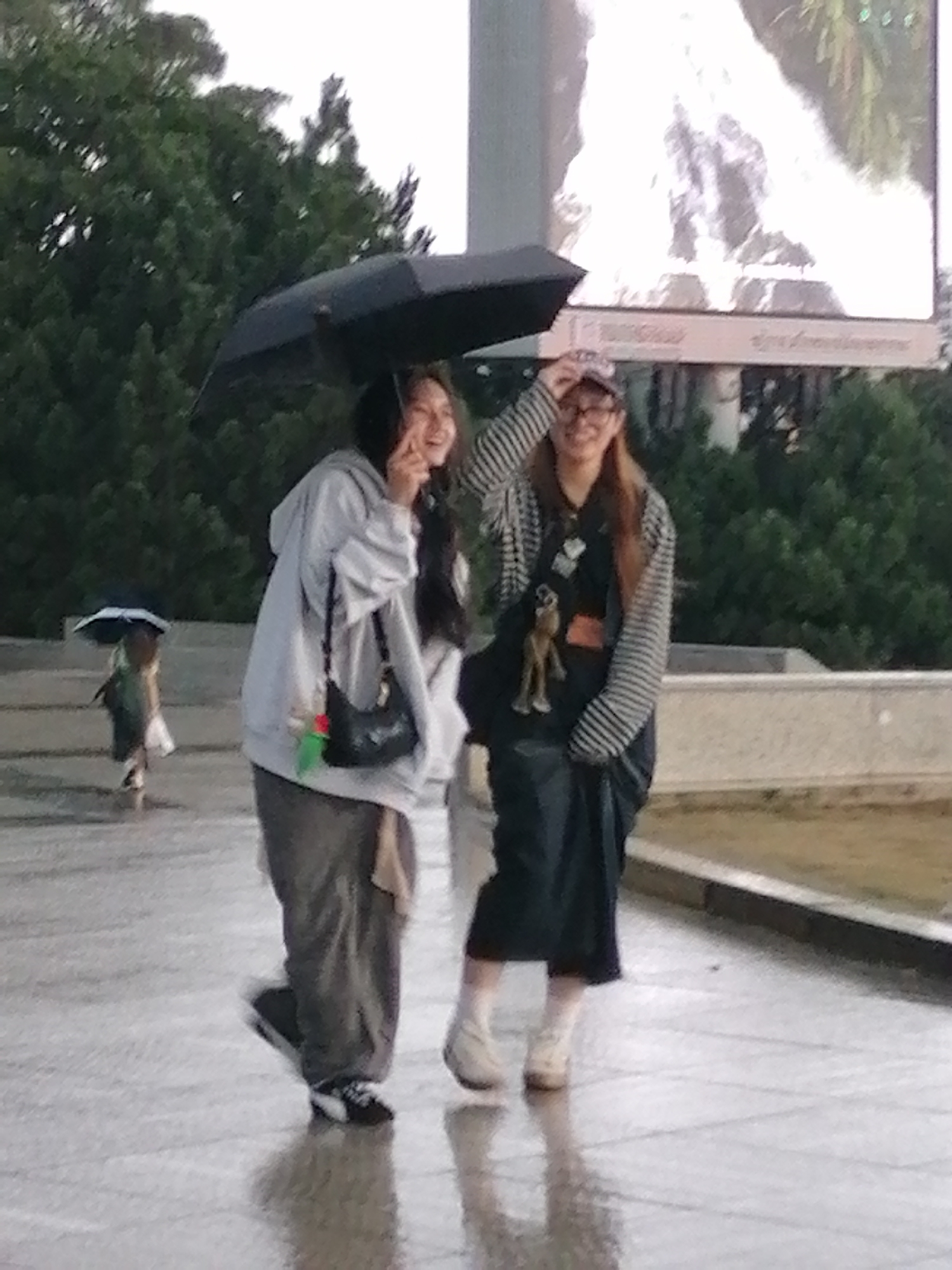 In Da Lat, Vietnam, two young women are walking in the rain. The woman on the left is wearing a light grey hooded sweatshirt, dark grey pants, and black and white sneakers. She is holding a black umbrella over her head and carrying a black shoulder bag with a green charm. The woman on the right is wearing a striped long-sleeved shirt, a dark skirt, and light-colored boots. She has glasses and a cap on her head and is holding her hand up to adjust it. A stuffed animal is attached to her bag. The ground is wet and reflective, indicating recent rainfall. In the background, there are trees and a large billboard displaying a bright image. Another person is visible in the distance, also under an umbrella. The scene appears to be an outdoor public space, possibly a park or pedestrian walkway.