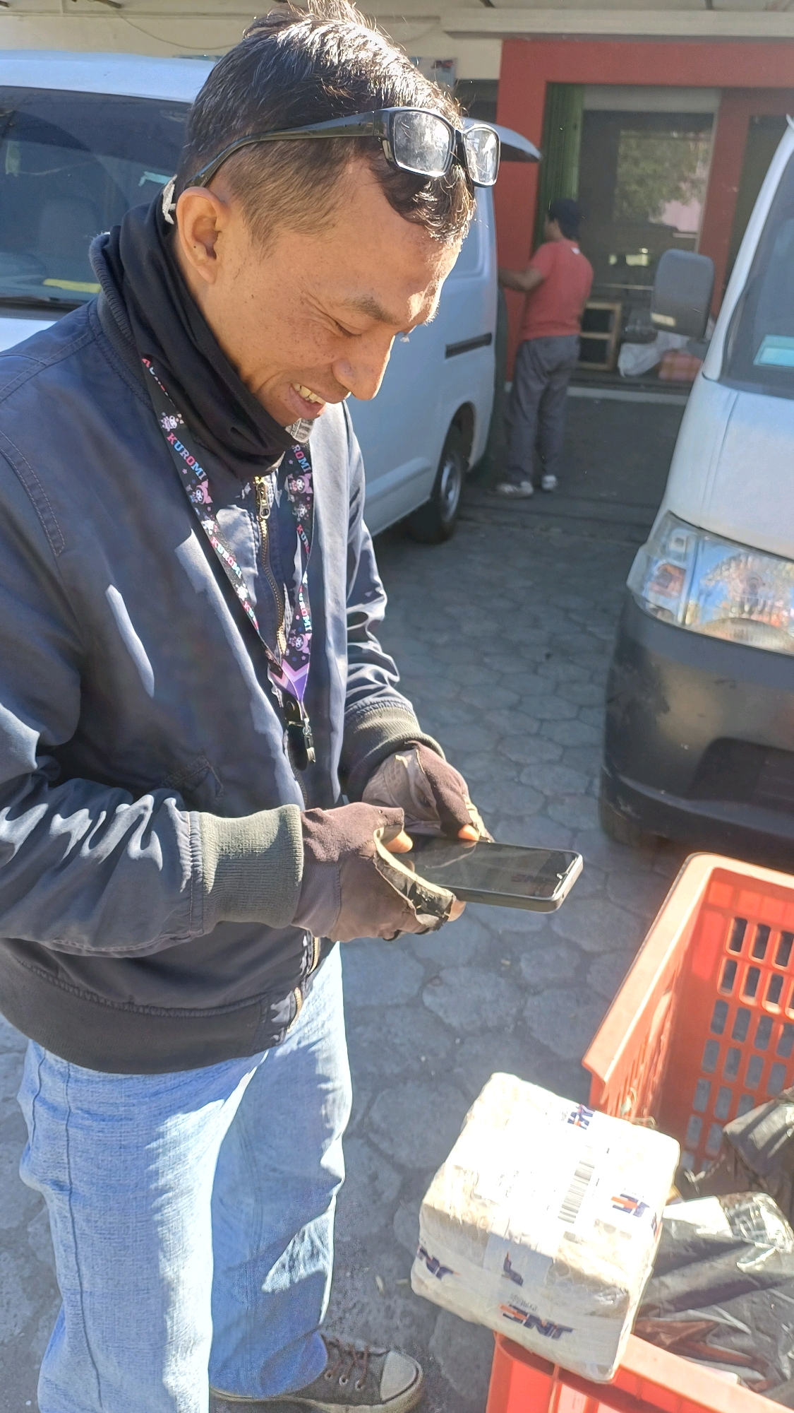 An outdoor, medium shot captures a man of Asian descent, wearing glasses atop his head, a dark jacket, light blue jeans, and fingerless gloves, as he smiles and looks down at a smartphone held in his hands. He is positioned in front of a white van, and to his right, a red plastic crate contains a wrapped package. In the background, another white van and a red building are visible. The scene is set on a paved surface, suggesting an urban or commercial area, possibly a delivery hub or a street in Kedu, Indonesia. The lighting indicates a sunny day.